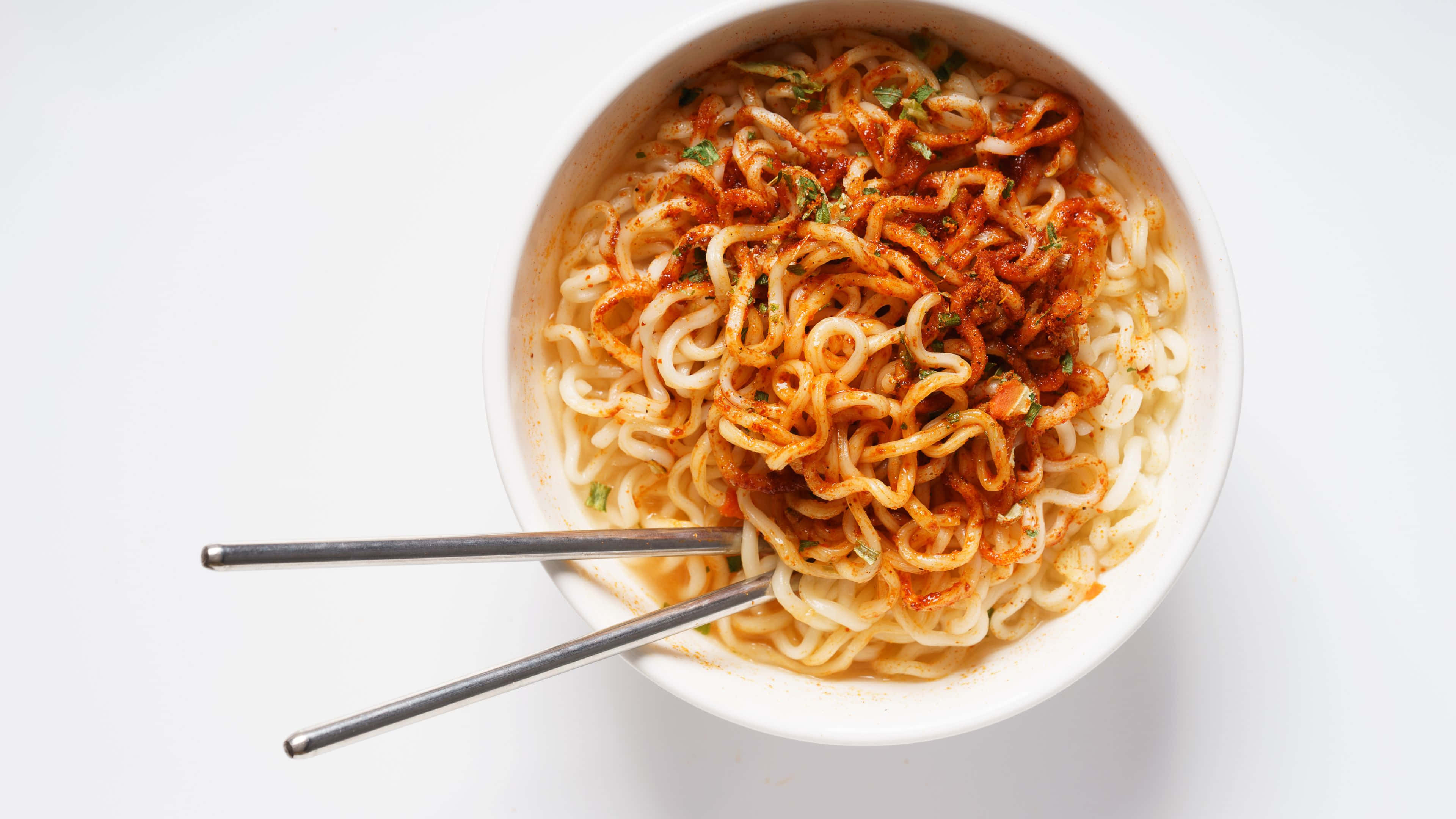 Noodles 桌布 壁紙 — 俯視一個白色碗，裡面裝滿辛辣拉麵，上面有紅色調味料和綠色香草，旁邊放著筷子。