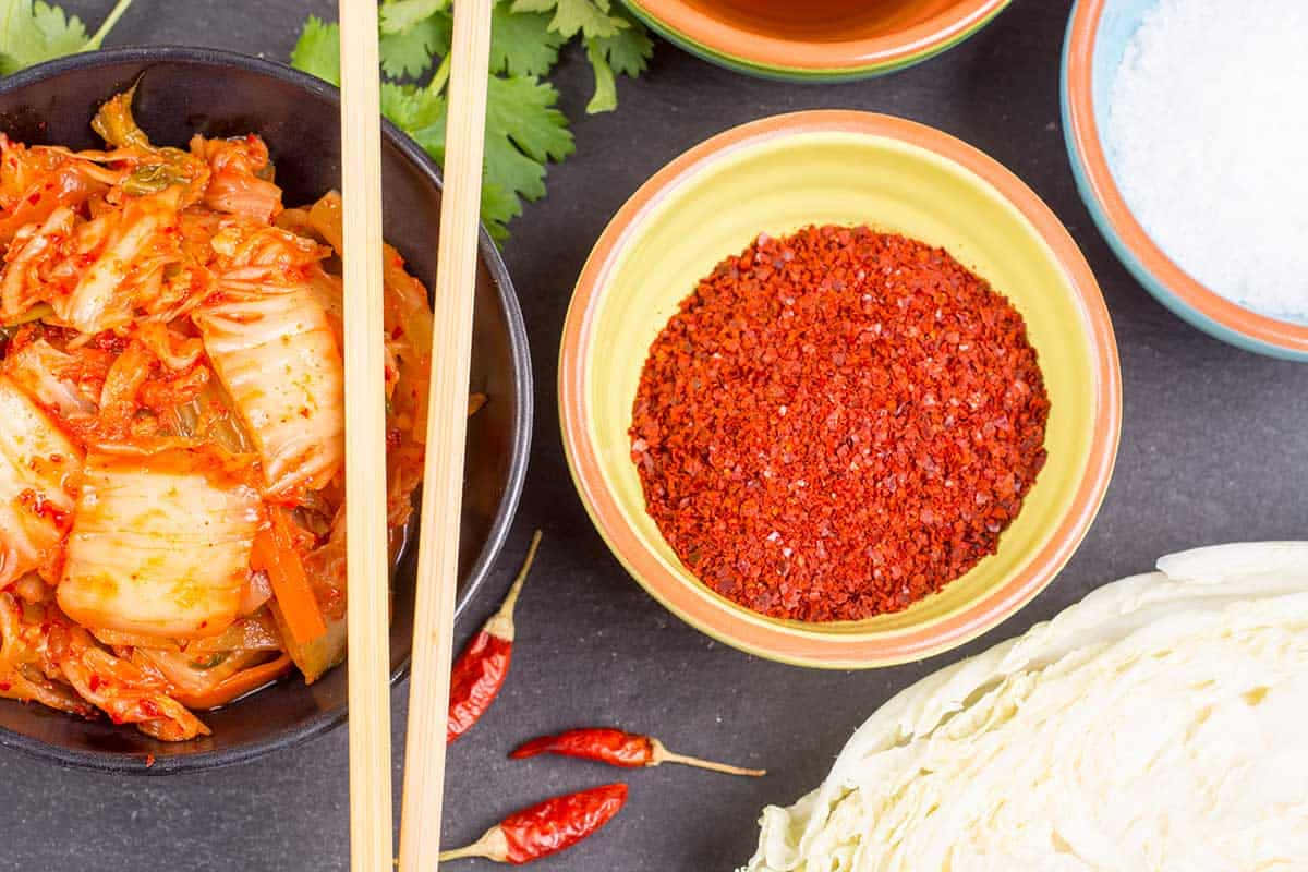 Red Chili Powder 桌布 壁紙 — 俯視圖，碗中盛有色彩鮮豔的泡菜，旁邊是紅辣椒片和新鮮食材，置於深色表面上。
