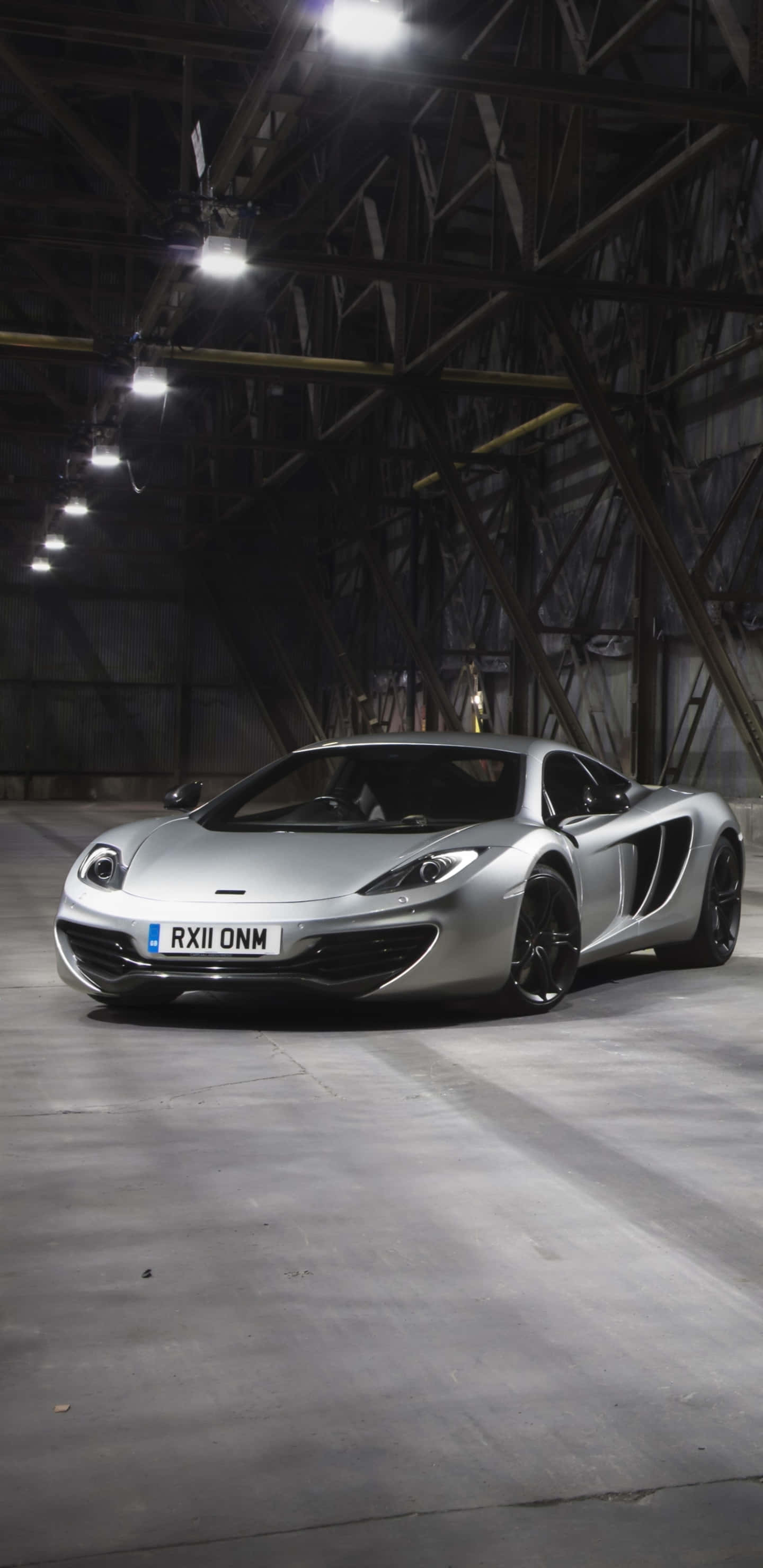 倉庫拍攝麥拉倫720S壁紙（Pixel 3XL格式） 壁紙 — 一架時尚的銀色McLaren跑車在寬敞倉庫內的工業燈光下閃閃發光，倉庫有外露的鋼樑。