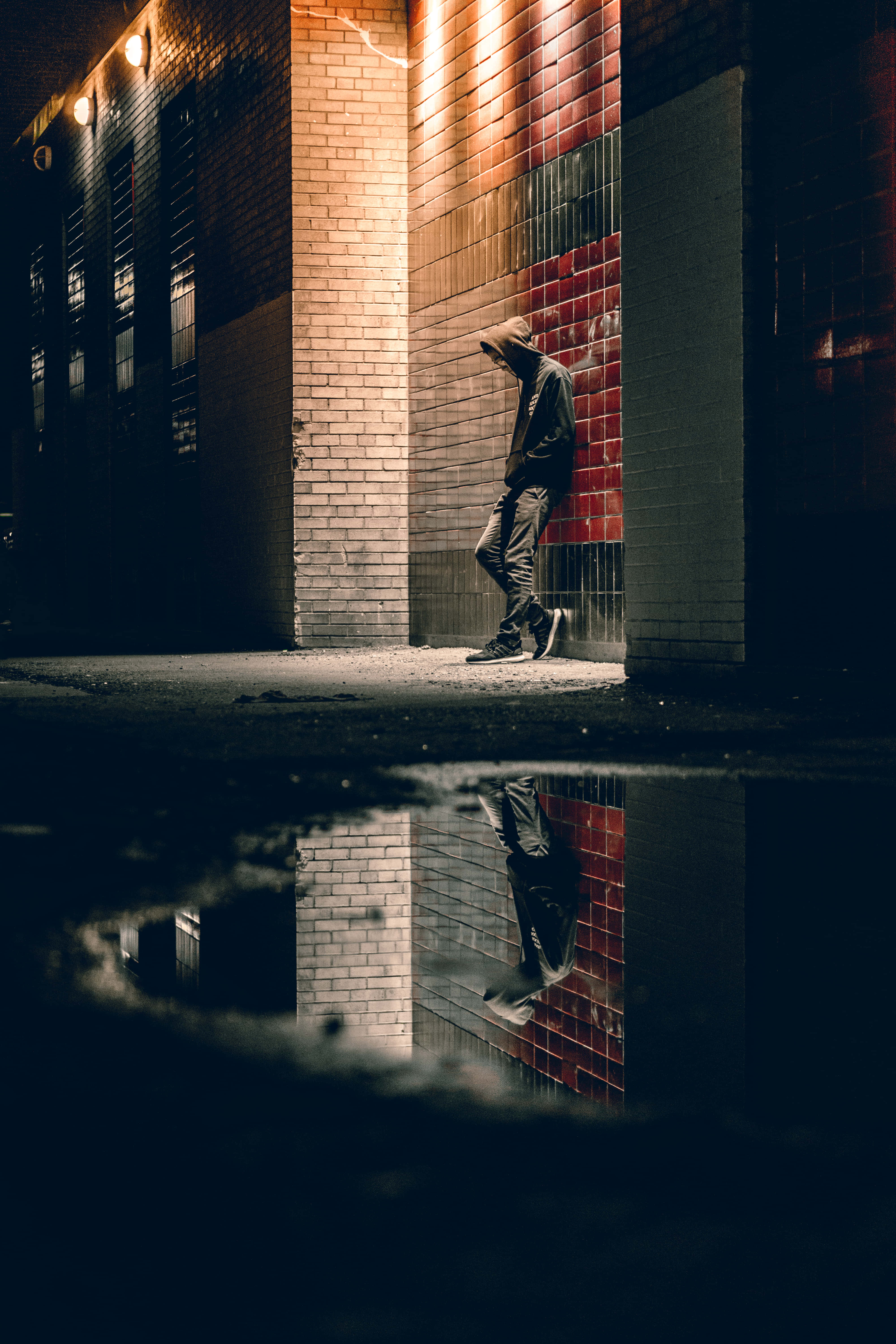 倒影中的悲傷男子 壁紙 — 夜晚，一個孤獨的戴帽人影靠在磚牆上，他們的倒影映照在下面的雨水坑中。