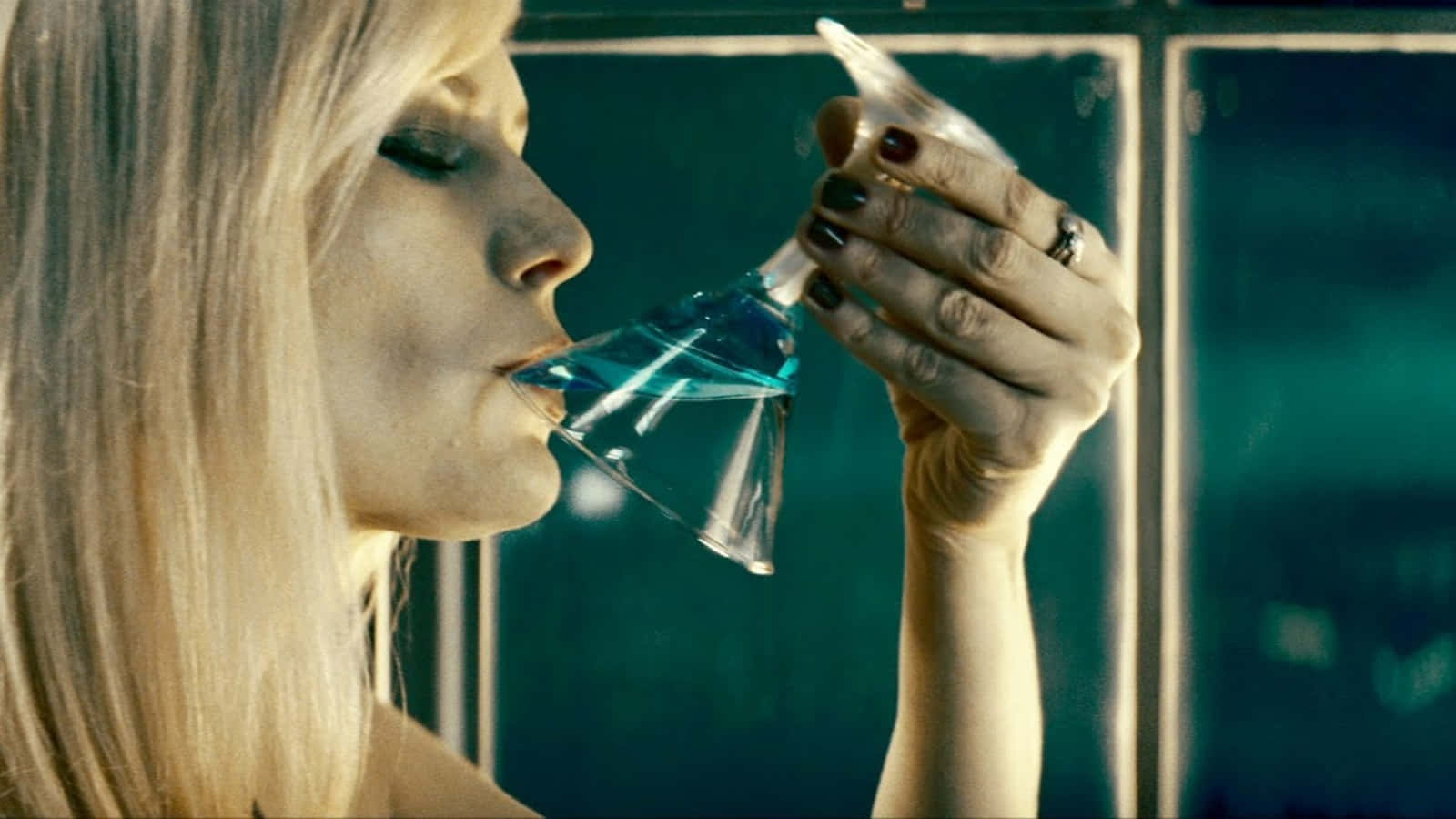 倒置視角下的壯麗秋日風景 壁紙 — 一位金髮女郎在燈光昏暗、牆壁呈青綠色的房間裡，從馬丁尼杯中啜飲藍色液體。