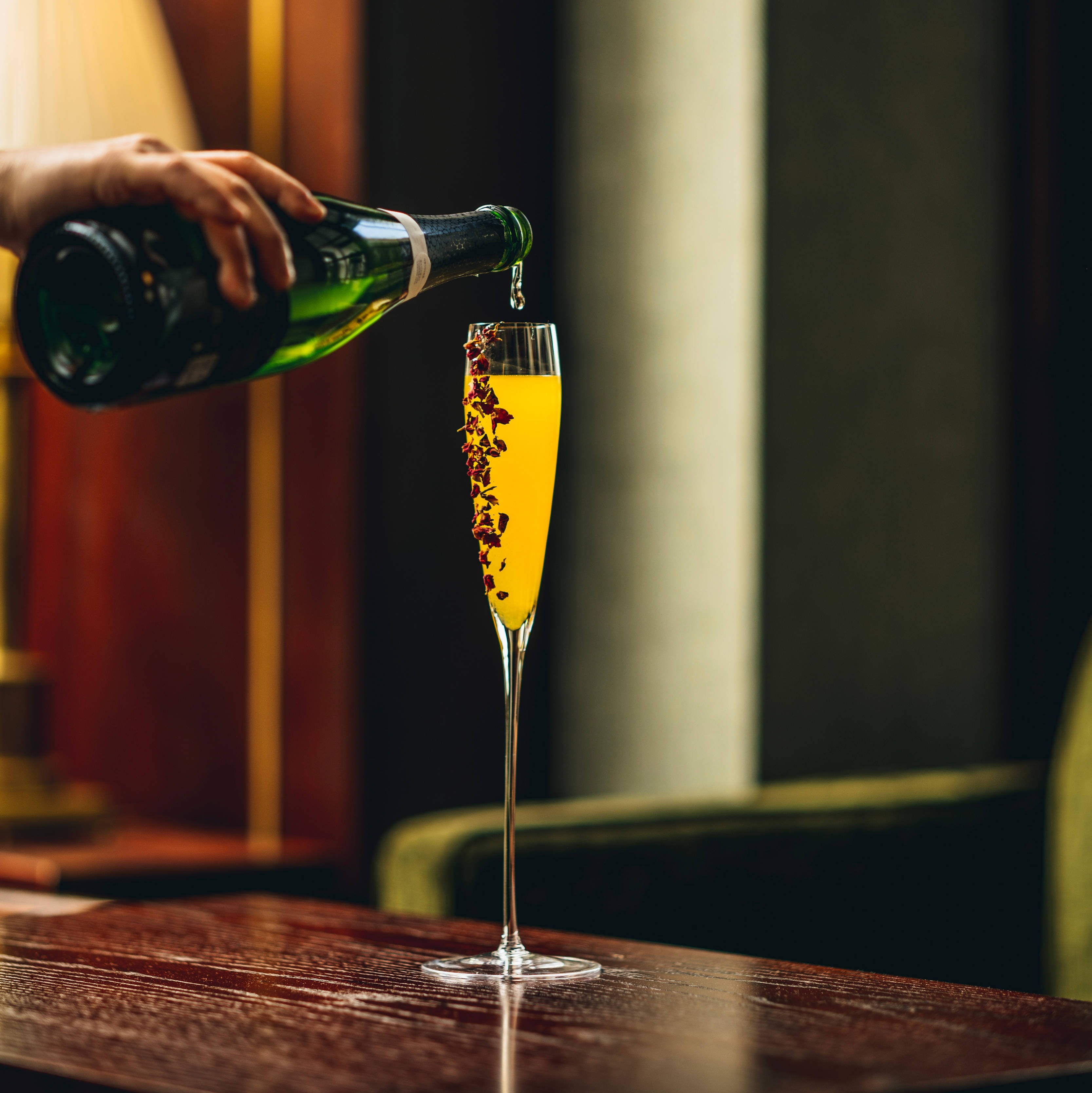 倒飲料 壁紙 — 一隻手將金色香檳倒入飾有乾玫瑰花瓣的笛形杯中，放在拋光木吧檯上。