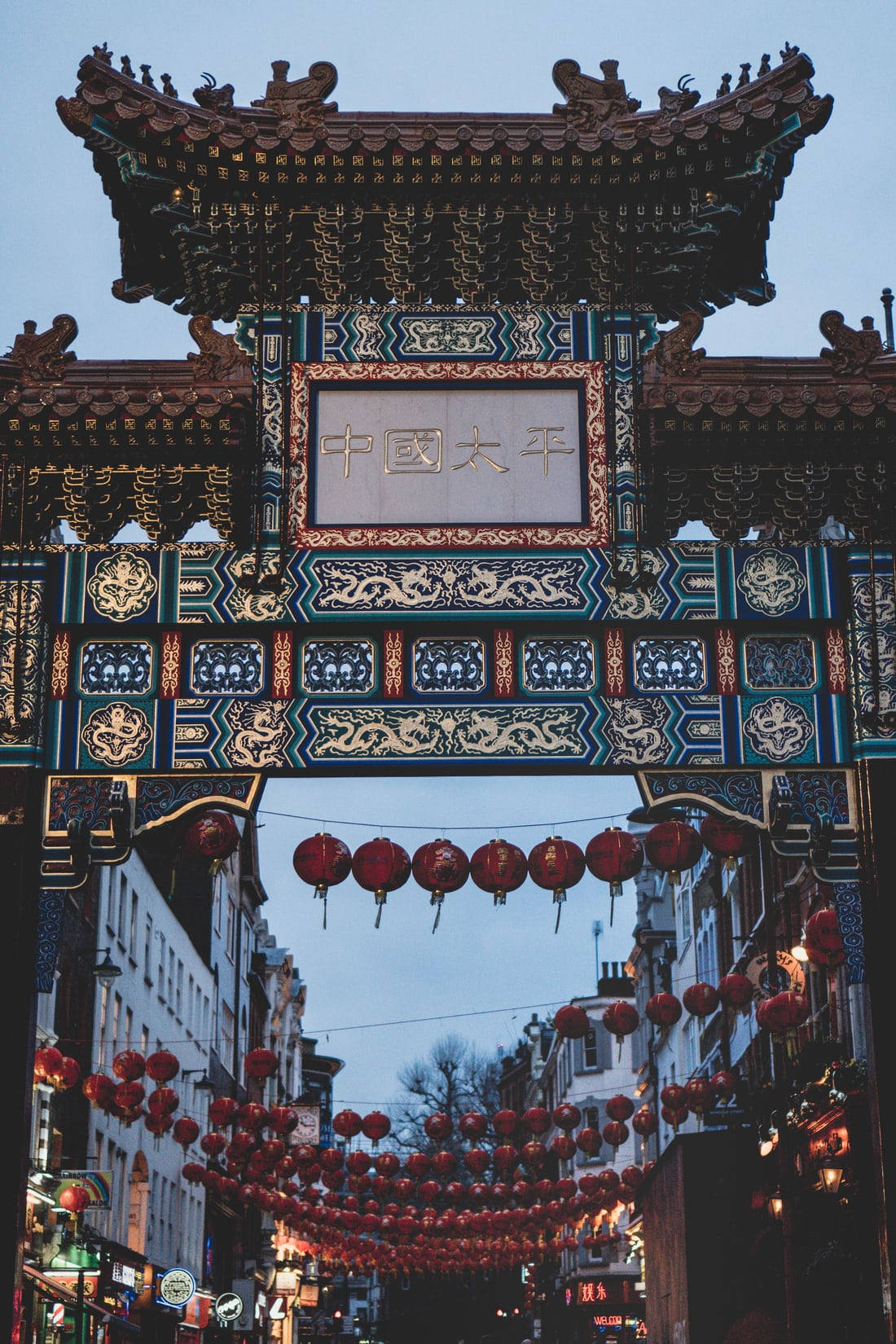 倫敦唐人街大門 壁紙 — 一個裝飾華麗的傳統中式牌坊，掛著紅燈籠，懸在黃昏時分的狹窄街道上。