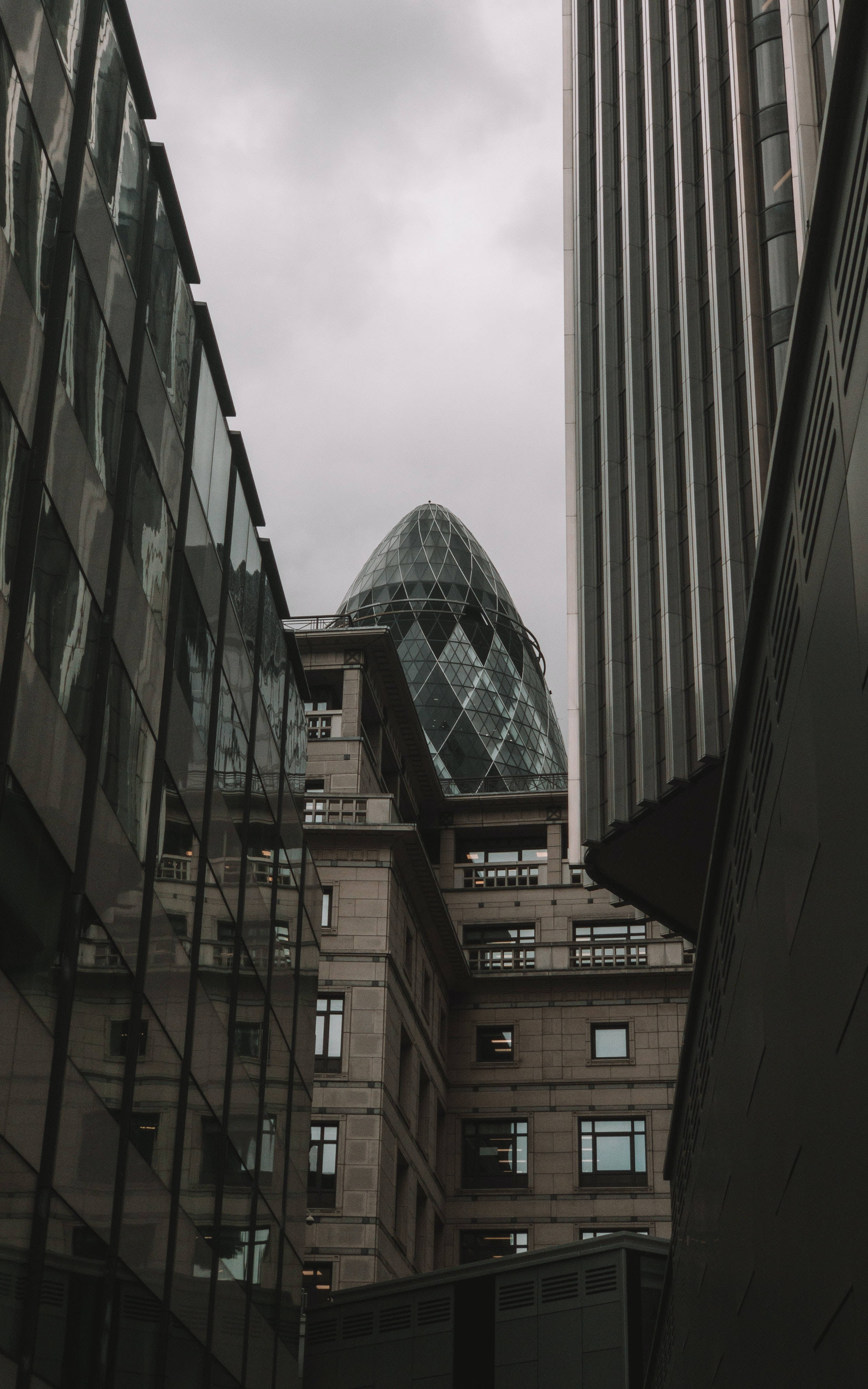 倫敦摩天大樓間的「小黄瓜」建築 壁紙 — 低角度觀看Gherkin摩天大樓在狹窄城市建築之間窺視，天空多雲。