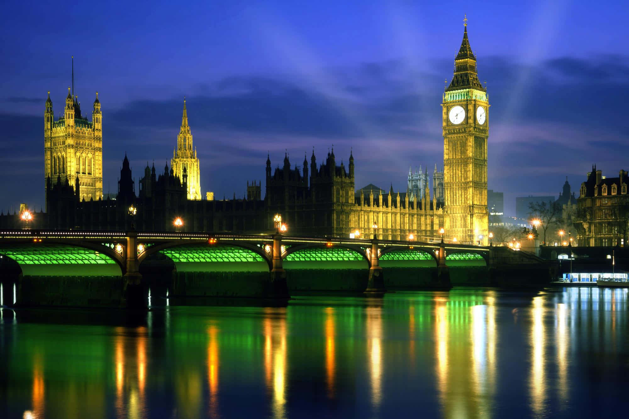 England 桌布 壁紙 — 倫敦標誌性的大笨鐘和國會大廈在黃昏時亮起，鮮綠色的橋樑燈光倒映在泰晤士河上。