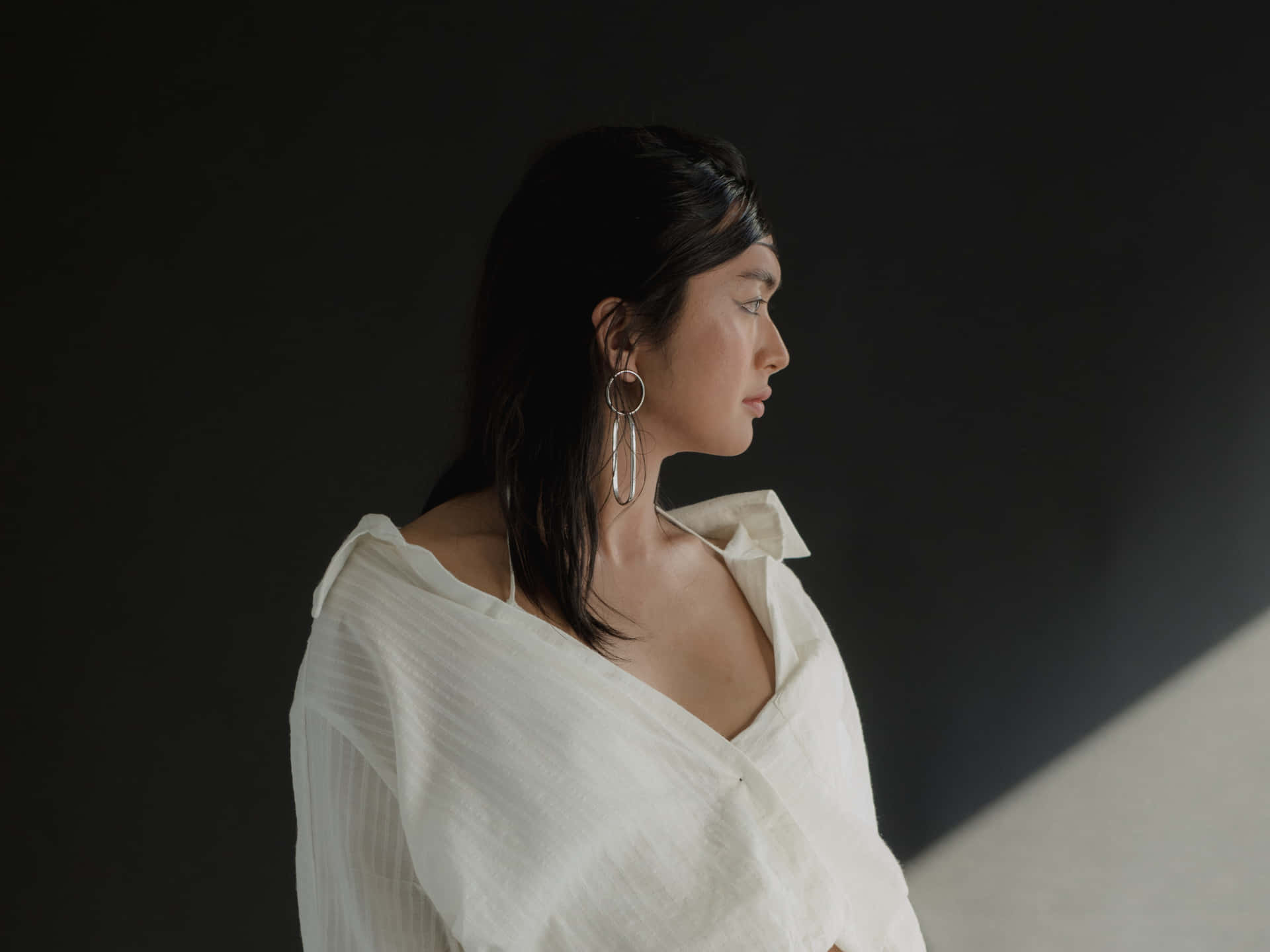 身穿白色襯衫、戴著大耳環的女性在昏暗的攝影棚燈光下若有所思地看向一旁。