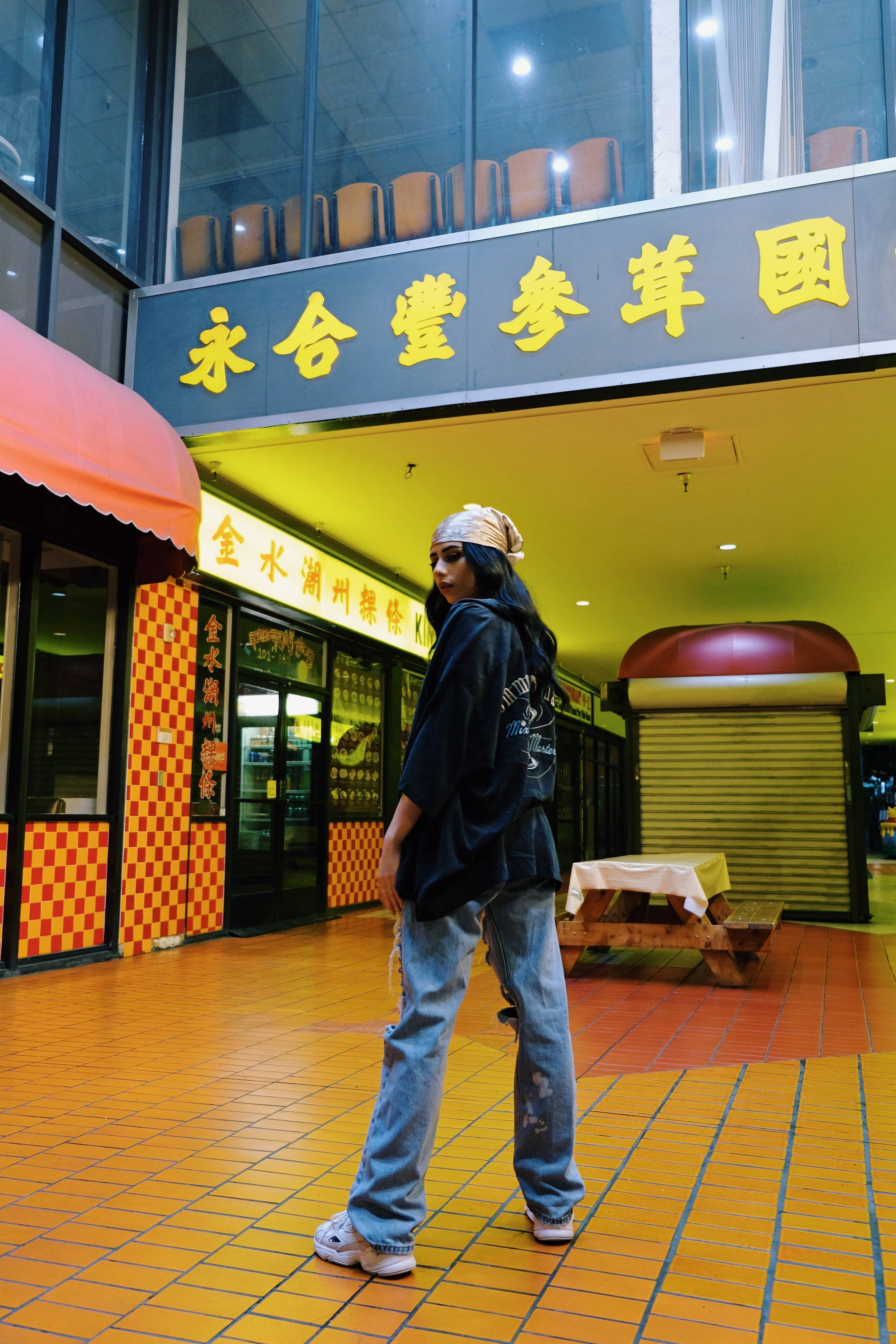 假小子逛中國商場 壁紙 — 一個戴著頭巾和超大外套的人走過一個光線充足的室內美食廣場，地板鋪有黃色瓷磚，並有中文標誌。