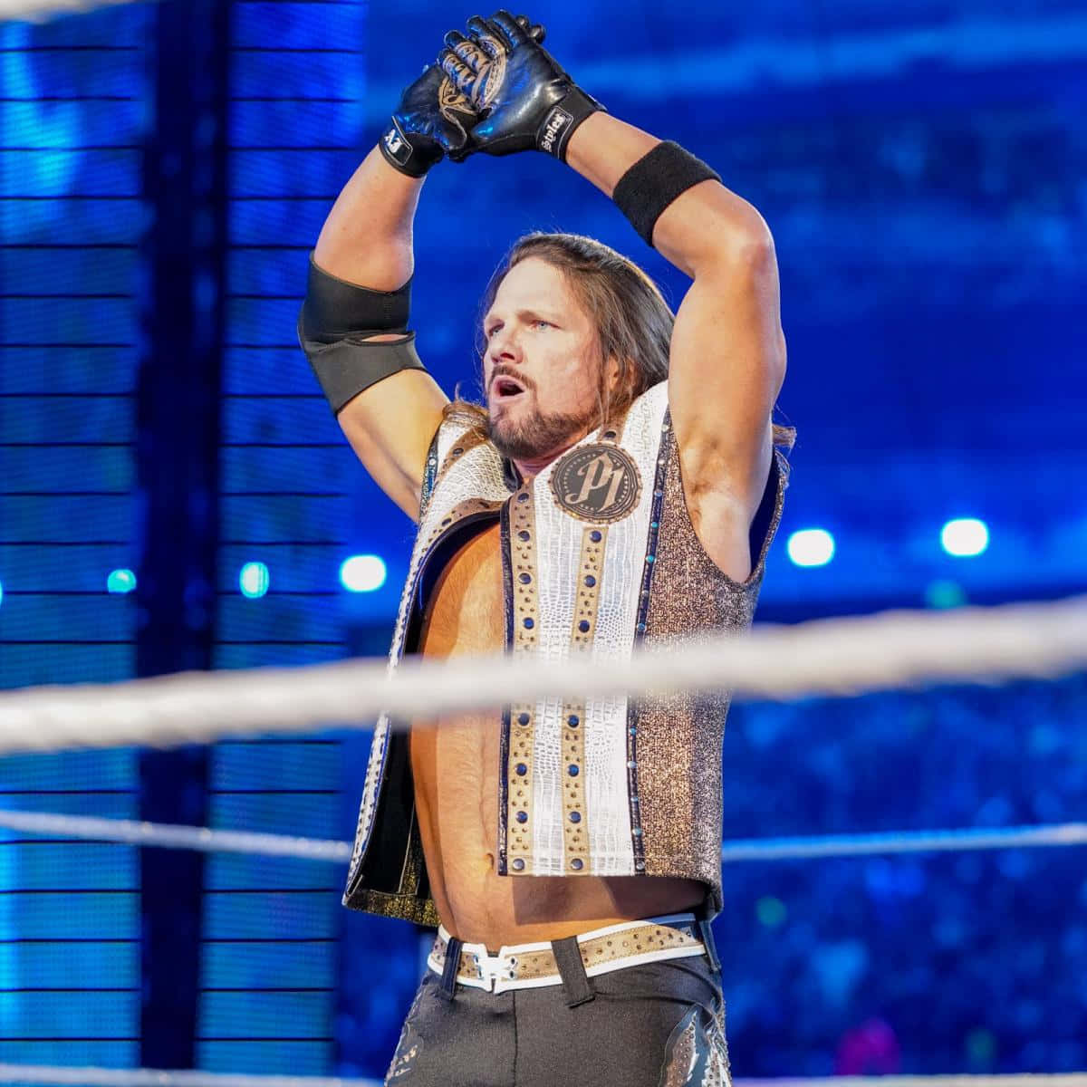 偉大的美國摔角手 AJ Styles 壁紙 — 一位肌肉發達的摔角手穿著閃閃發光的背心，在燈光明亮的擂台上舉起戴著手套的雙手慶祝勝利。