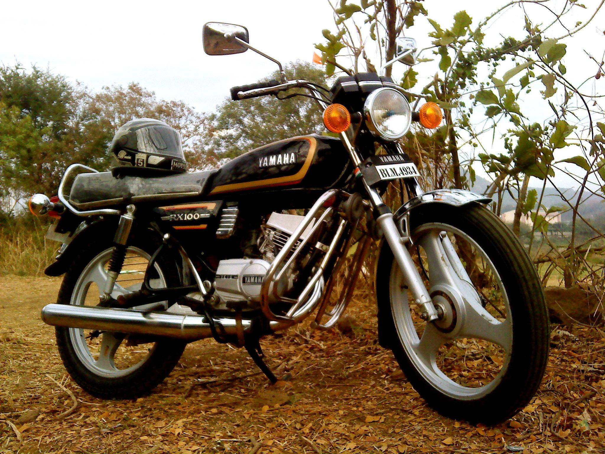 停在庭院中的黑色雅馬哈RX100摩托車 壁紙 — 一架經典黑色Yamaha RX100電單車停泊在乾葉上，旁邊有綠色灌木，天空多雲。