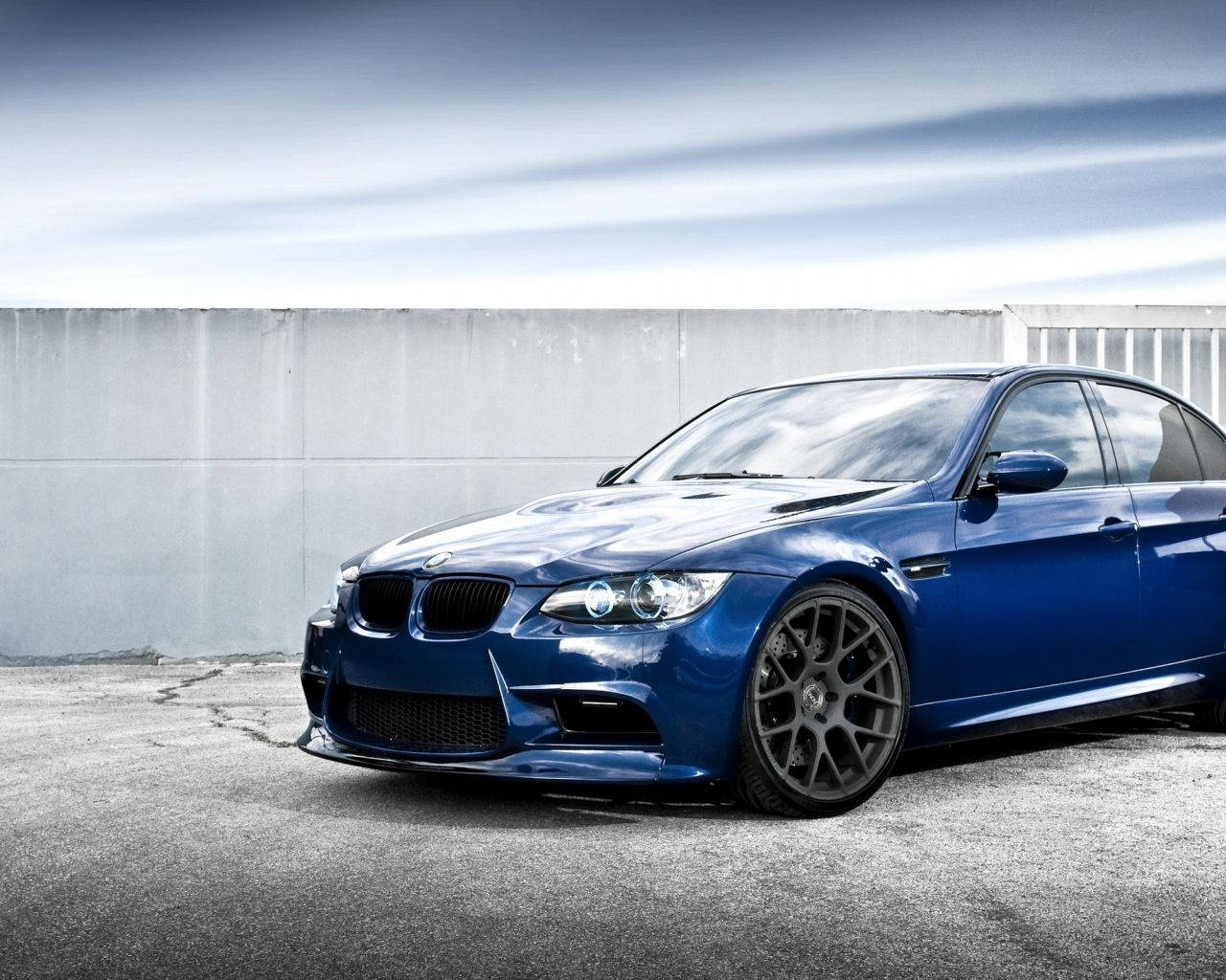 停泊於屋頂露台的藍色BMW 壁紙 — 一架時尚的藍色寶馬M3跑車停在裂開的瀝青路上，背景是簡約的混凝土牆，天空多雲。