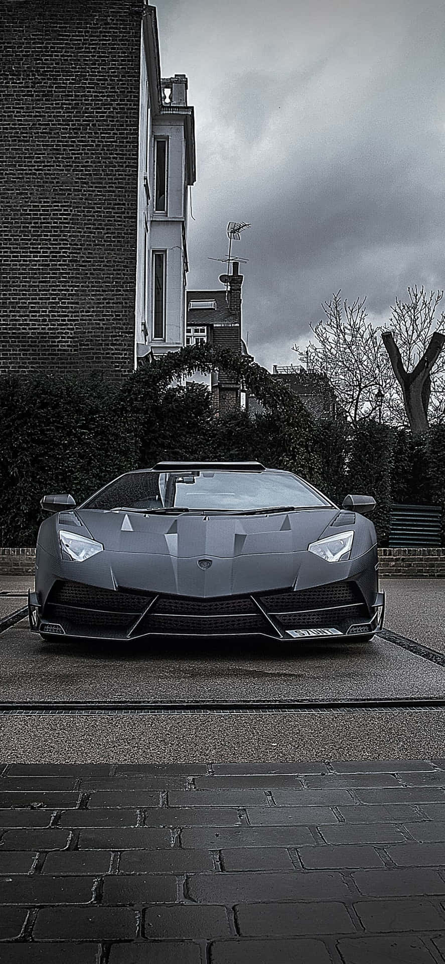 一輛灰色的林寶堅尼 Aventador 停泊在濕漉漉的鵝卵石街道上，天空多雲。