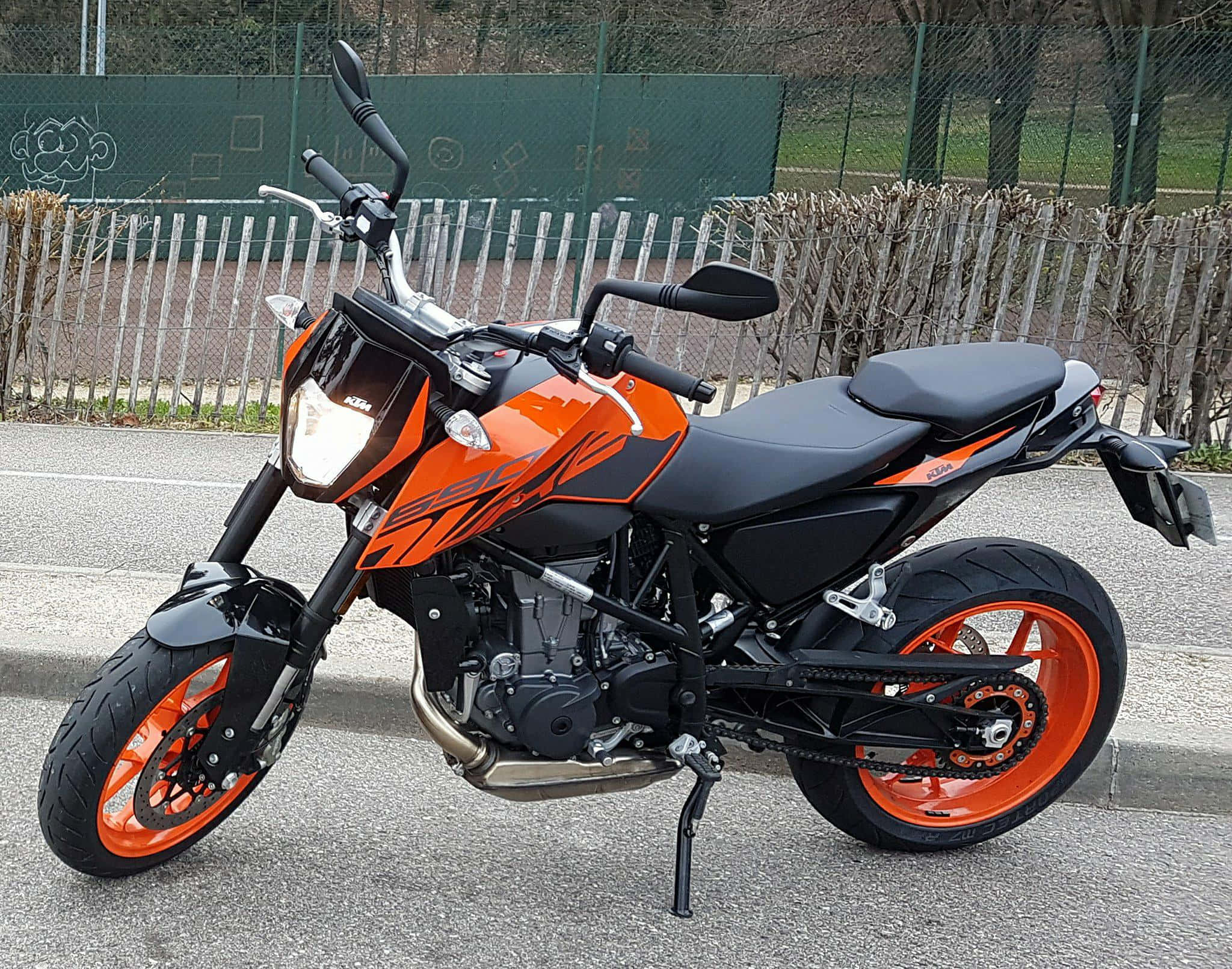 停泊的KTM Duke 690機車圖片 壁紙 — 一架搶眼的橙色和黑色KTM 390電單車停泊在鋪砌路面上，背景有圍欄和光禿禿的樹木。