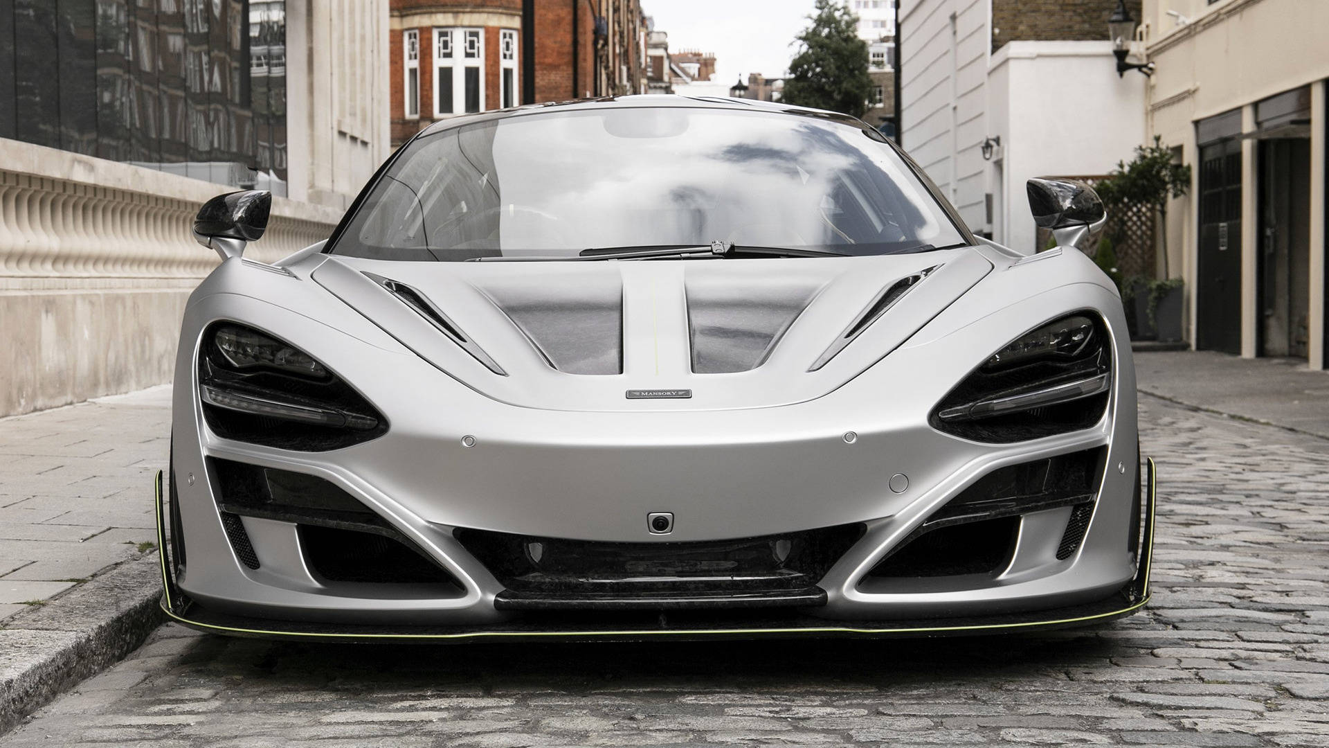 停泊的曼索里麥拉倫720S 4K 壁紙 — 一架時尚的銀色McLaren跑車，帶有黃色點綴，停泊在都市建築物之間的鵝卵石街道上。