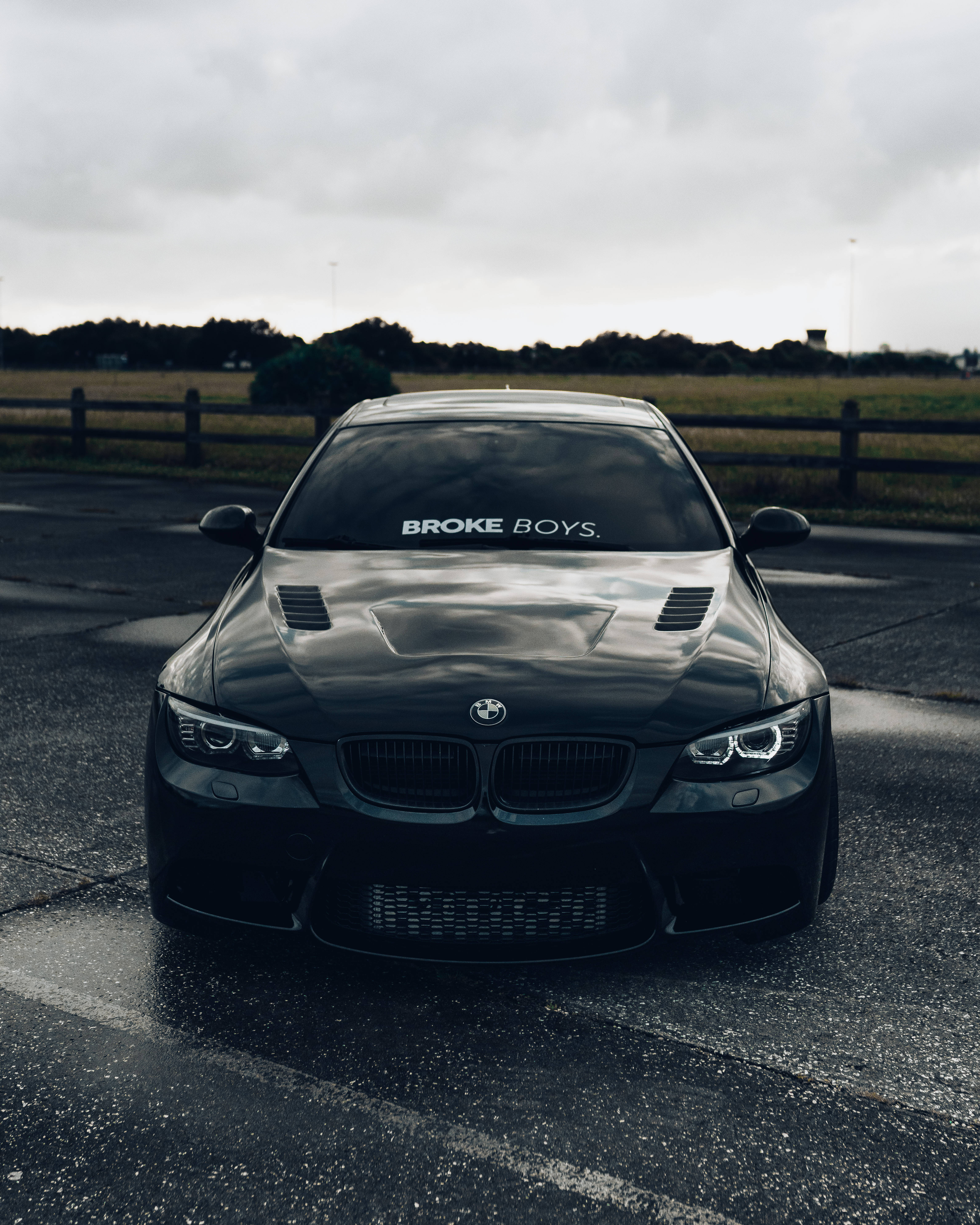 停泊的黑色寶馬 M3 壁紙 — 一架時尚黑色BMW跑車，擋風玻璃上貼有「BROKE BOYS」貼紙，停泊在潮濕的瀝青路上，天空多雲。