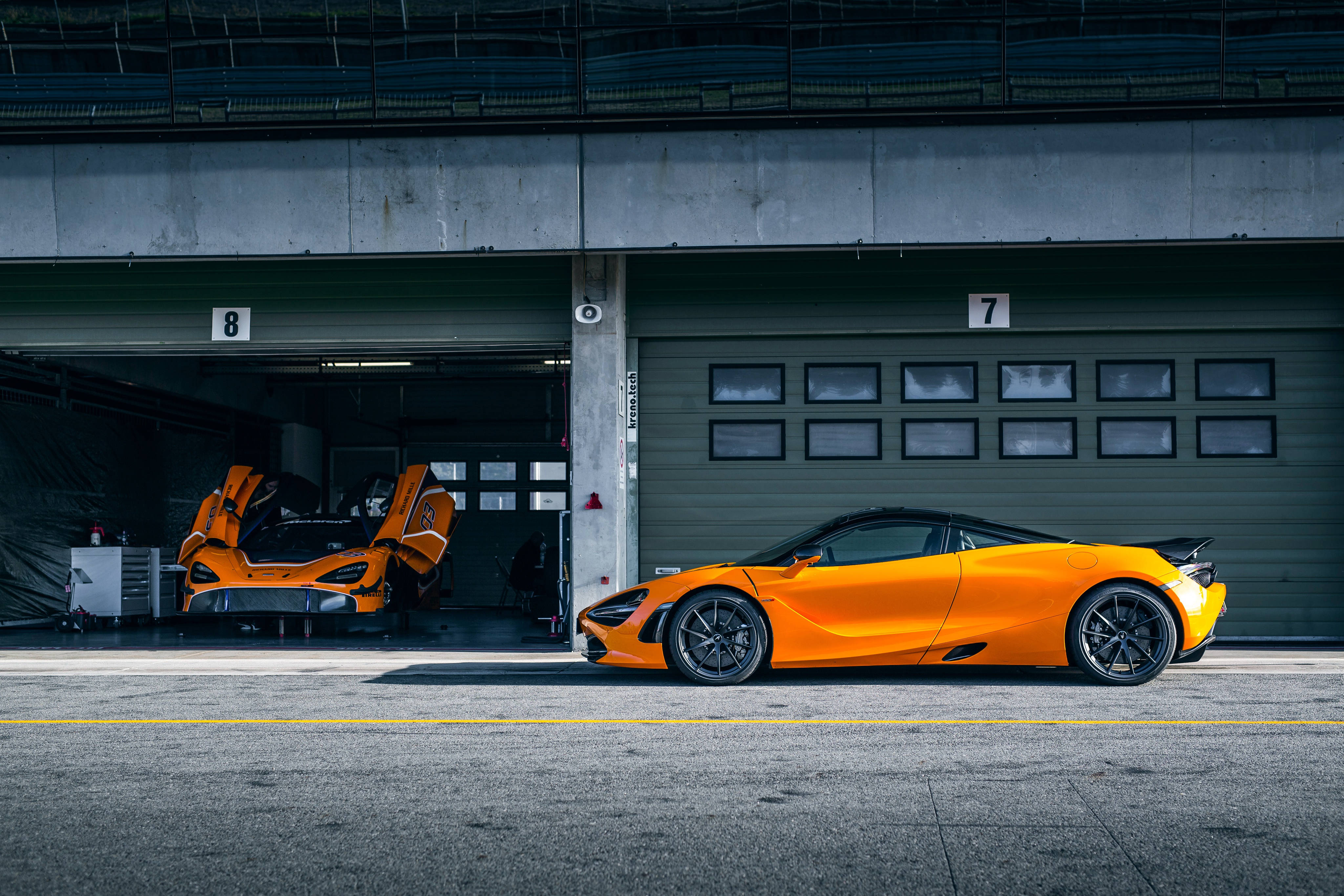 停靠於車庫旁的黃橙色麥拉倫720S 壁紙 — 兩輛充滿活力的橙色McLaren超級跑車停在混凝土車庫中，其中一扇車門打開，露出內部。