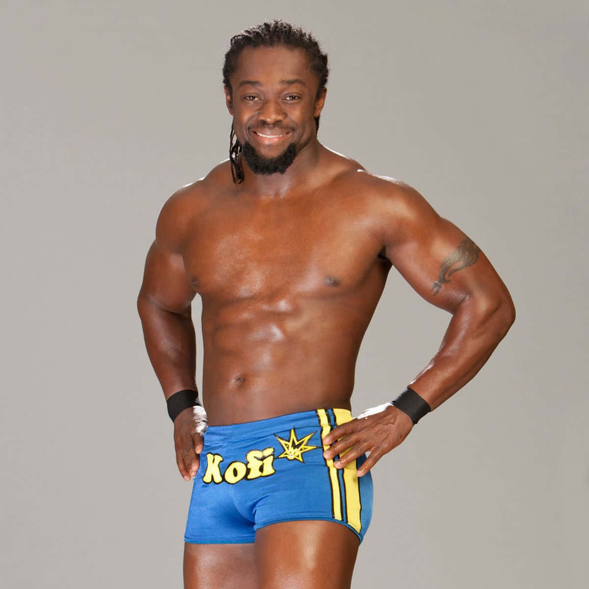 健壯的Kofi Kingston 壁紙 — 一位肌肉發達的摔角手穿著藍色和黃色短褲，雙手叉腰，在純灰色背景前自信地微笑。