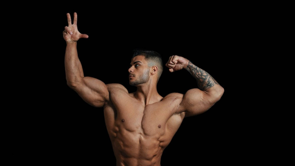 一個有紋身、赤裸上身的肌肉男在黑色背景前屈臂並做出和平手勢。