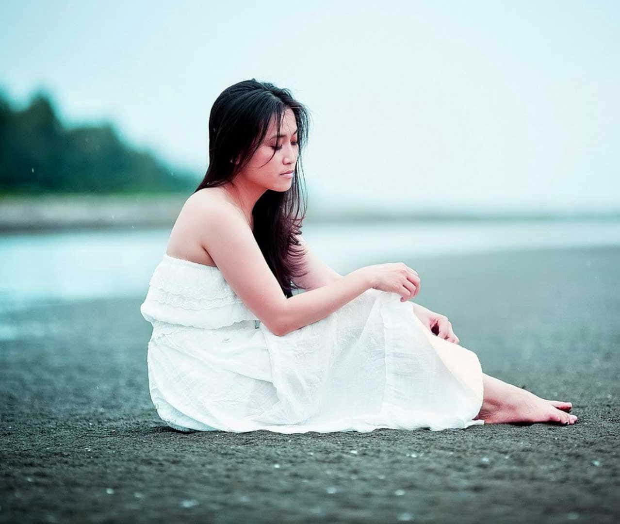 一位身穿白色連衣裙的女子在雨中坐在濕漉漉的海灘上，低頭帶著平靜的表情。