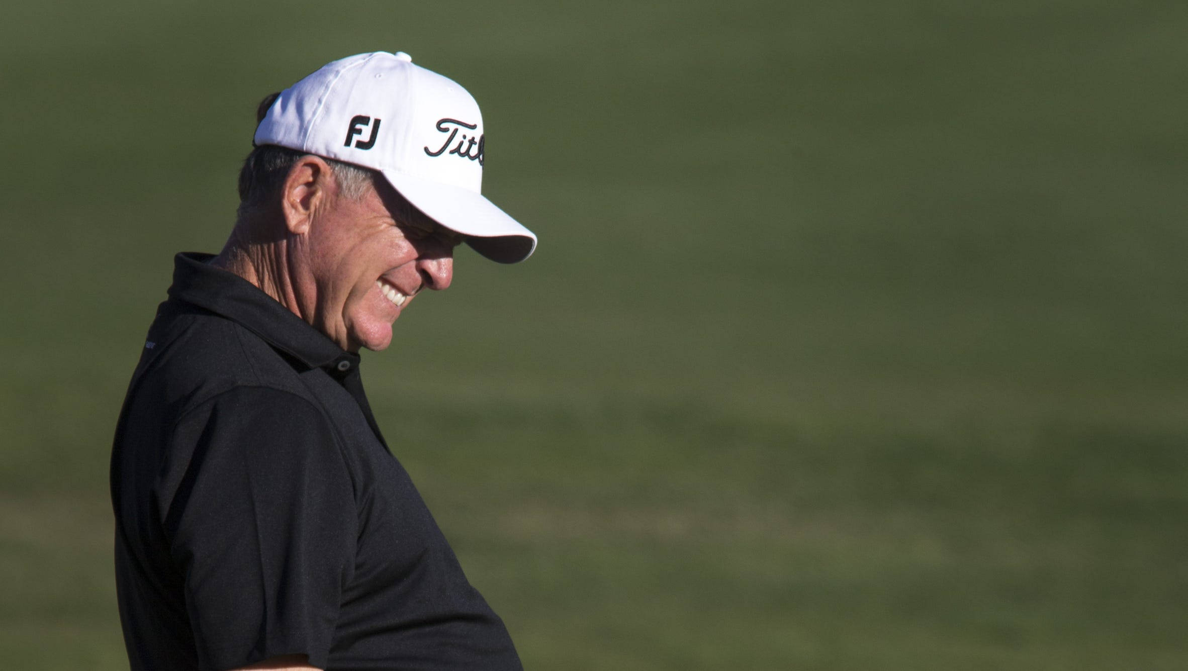 傑伊·哈斯在高爾夫賽事中展露笑容 壁紙 — 一位微笑的高爾夫球手戴著白色Titleist帽子和黑色Polo衫，在陽光明媚的綠色高爾夫球場上開懷大笑。