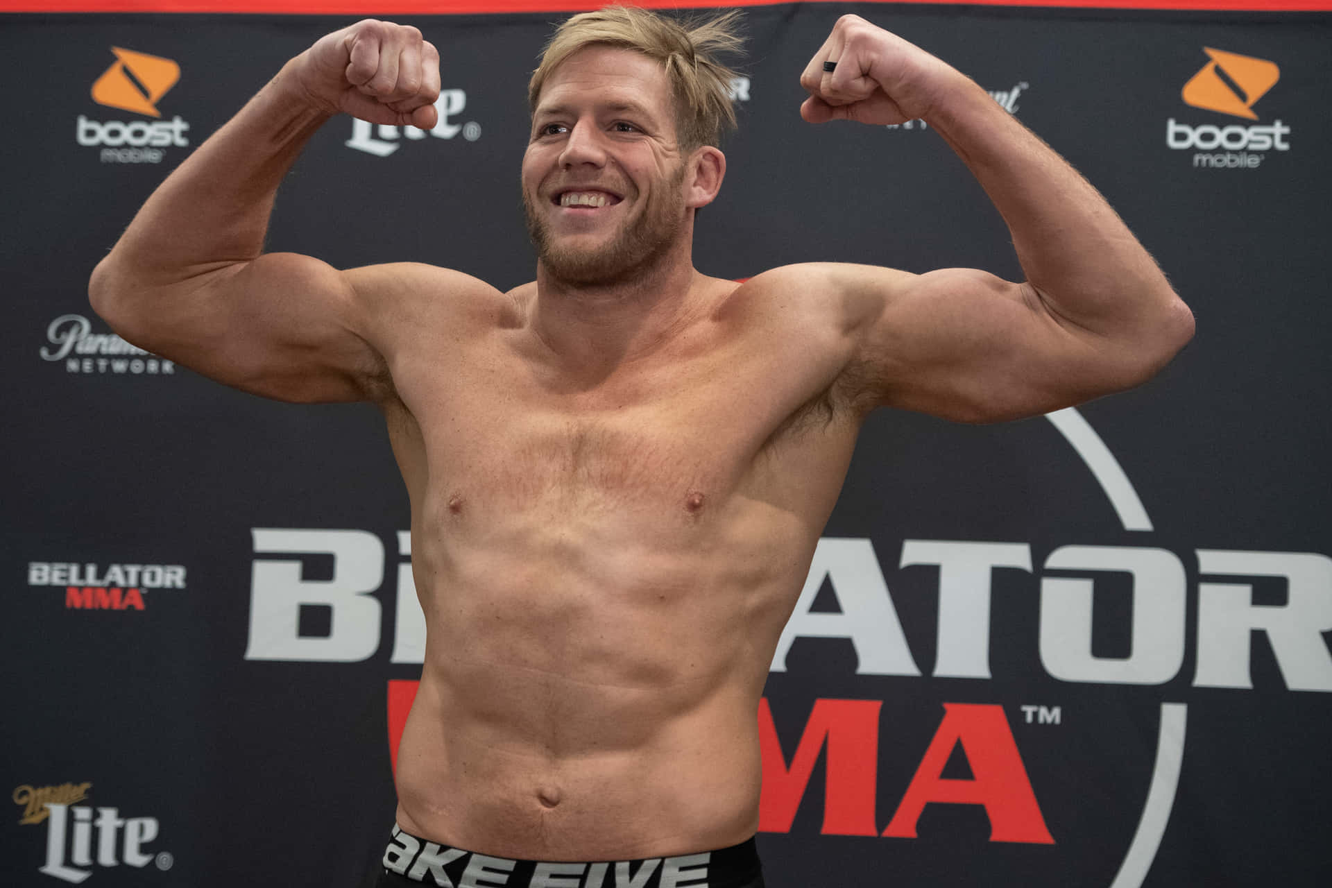 一位男性MMA拳手在稱重活動中展示肌肉並微笑。