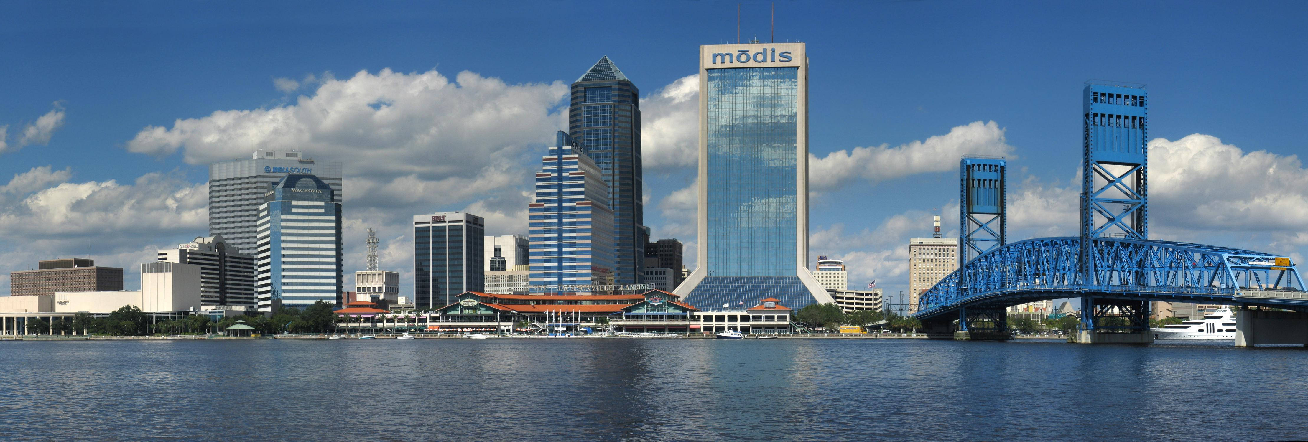 傑克遜維爾 Modis 大樓下午天際線 壁紙 — 現代城市天際線的全景視圖，玻璃摩天大樓和藍色橋樑橫跨平靜的水面，天空部分多雲。