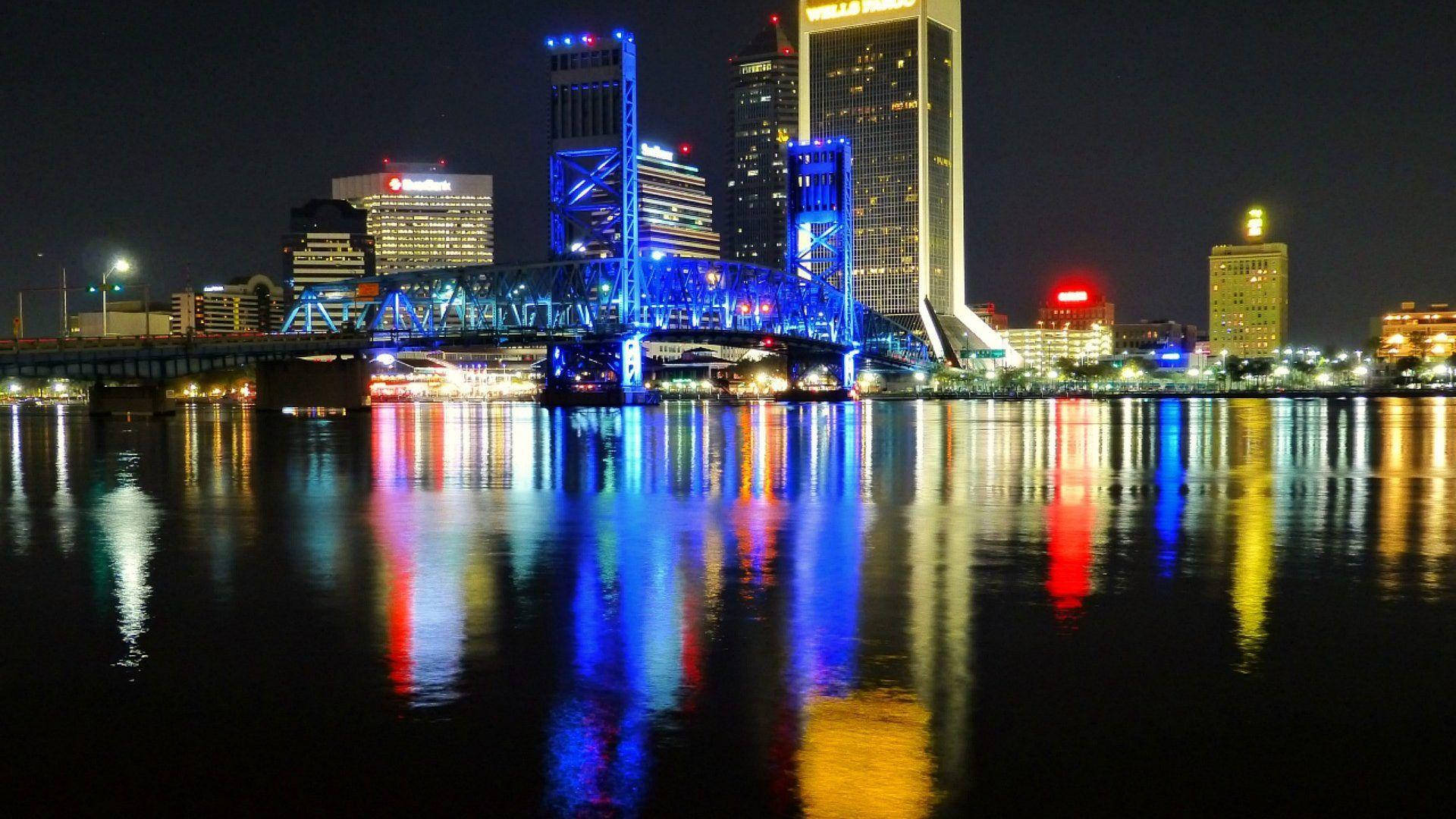 傑克遜維爾主街橋藍色燈光夜景 壁紙 — 充滿活力的夜間城市景觀，發光的藍色橋樑和摩天大樓倒映在平靜的水面上。