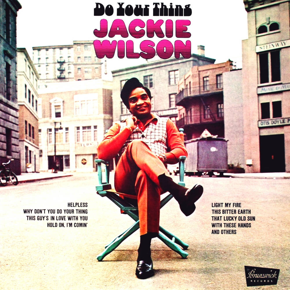 傑基·威爾遜美國歌手表演者 壁紙 — Jackie Wilson 自信地微笑，坐在導演椅上，身處陽光明媚的城市街道，周圍有復古建築和專輯文字。