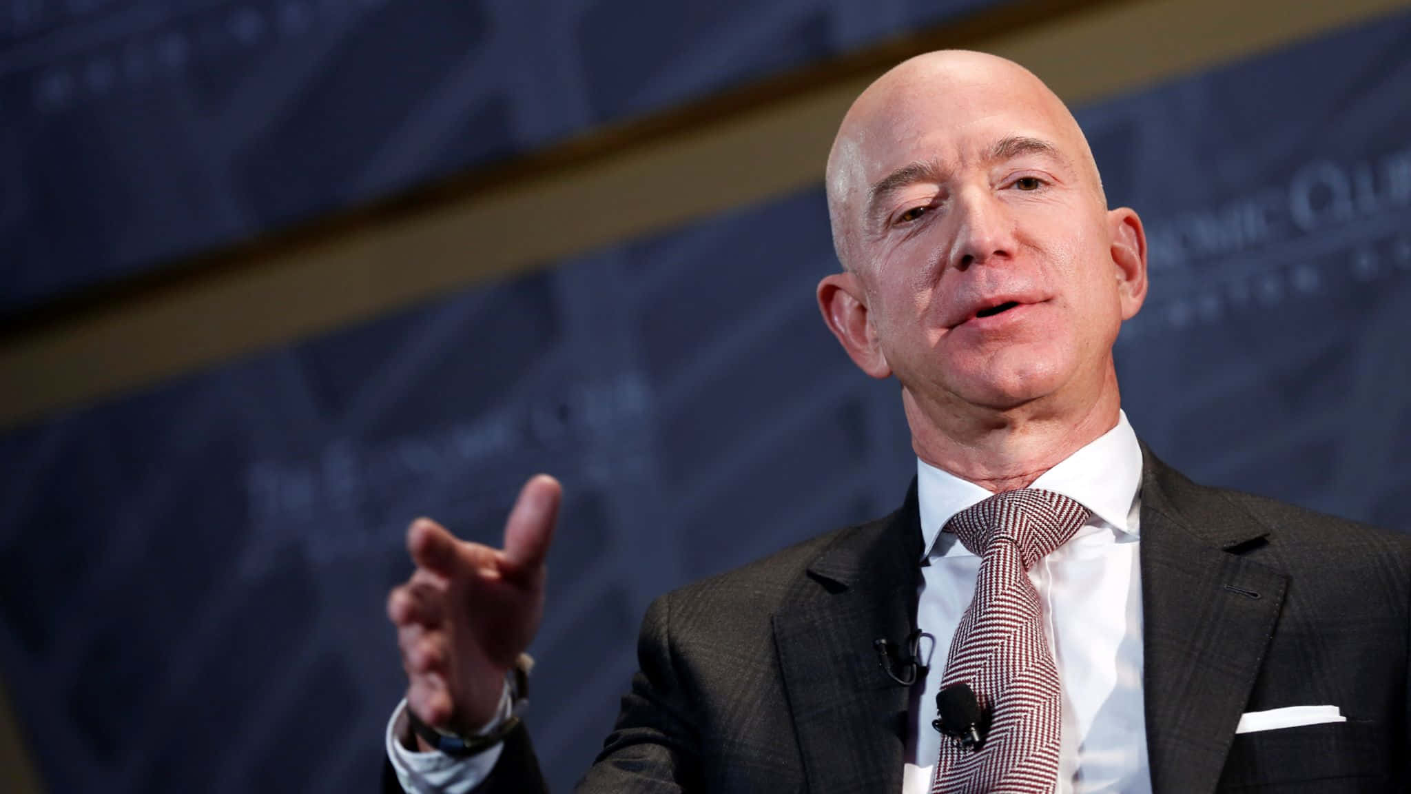 Jeff Bezos 桌布 壁紙 — 傑夫·貝索斯在正式活動中發言，身穿深色西裝和圖案領帶，背景為模糊的藍色。