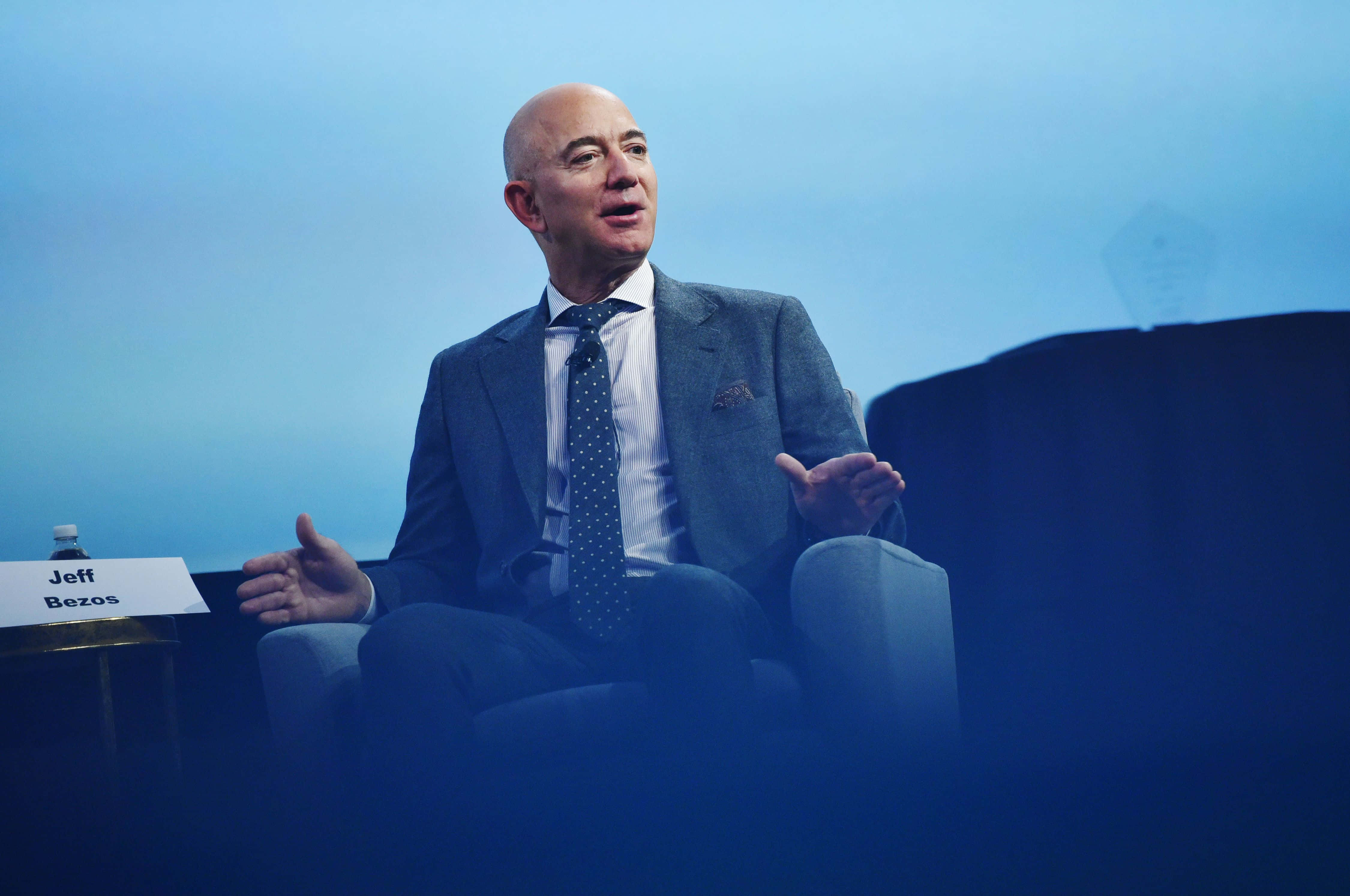 Jeff Bezos 桌布 壁紙 — 傑夫·貝索斯在藍色燈光的會議室中，穿著灰色西裝和波點領帶，生動地說話。