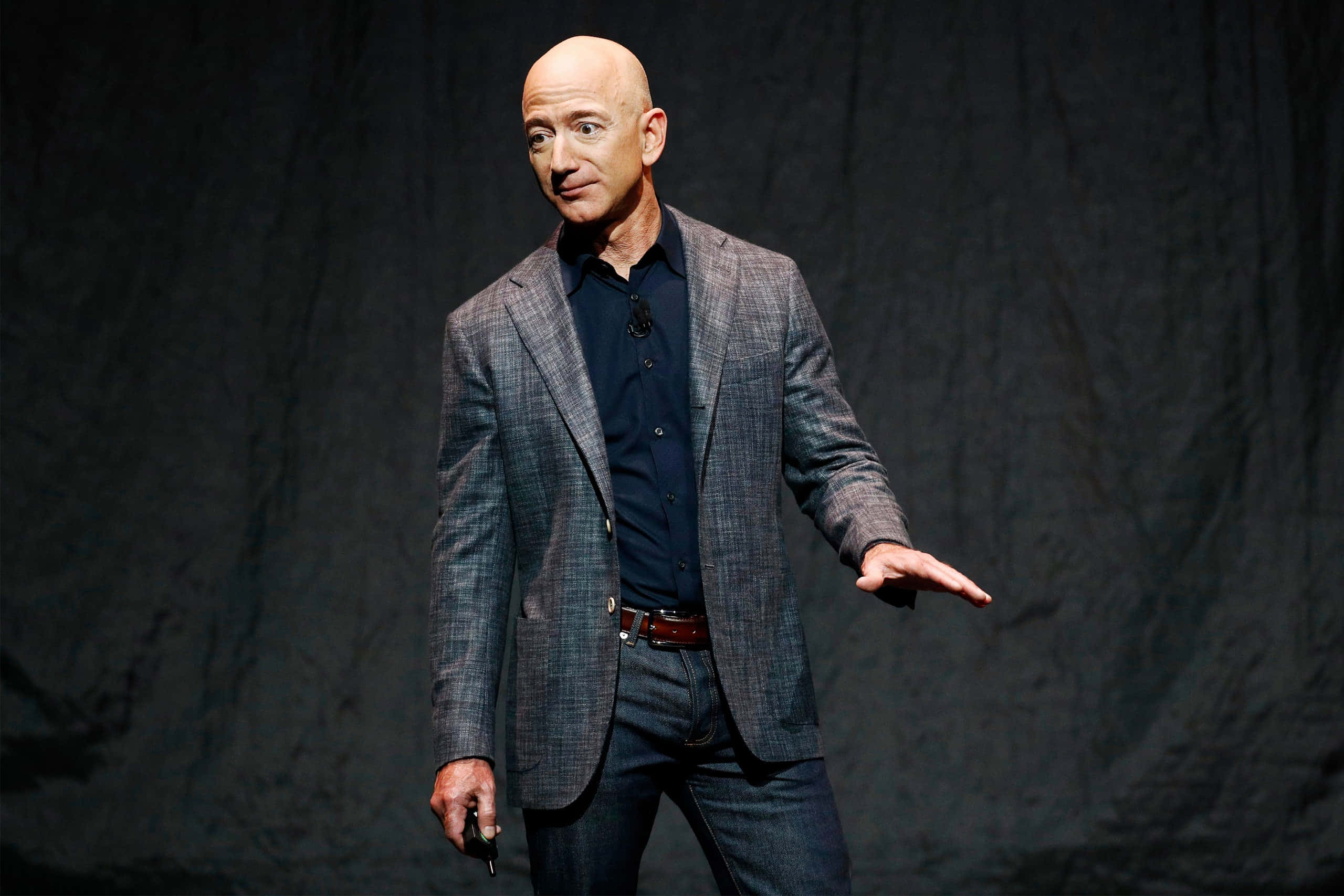 Jeff Bezos 桌布 壁紙 — 傑夫·貝索斯自信地站在舞台上，身穿格紋西裝外套，背景昏暗，在明亮的聚光燈下演講時做手勢。
