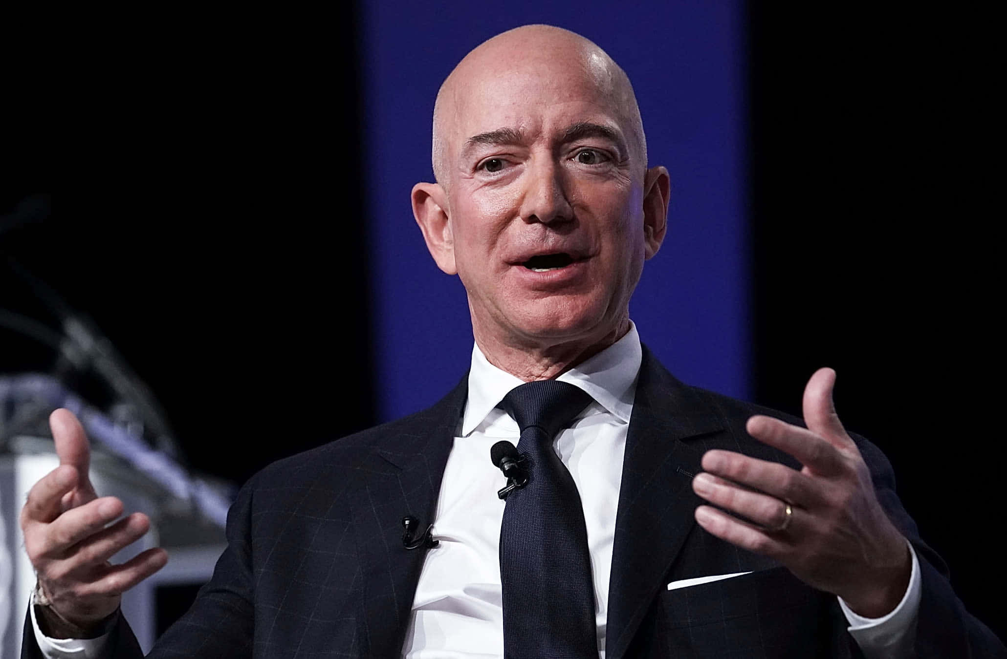 Jeff Bezos 桌布 壁紙 — 傑夫·貝索斯身穿深色西裝，在藍色背景前，雙手在句子中間做手勢，充滿激情地演講。