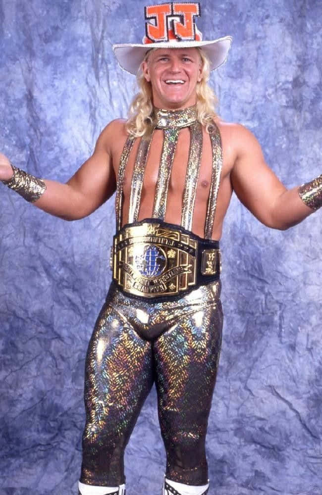 傑夫·賈雷特牛仔服裝 WWE 摔角手 壁紙 — 一位微笑的摔角手，身穿閃閃發光的金色和黑色服裝，腰間繫著冠軍腰帶，頭戴白色帽子，帽上有橙色字母，在帶有紋理的藍色背景前自信地擺姿勢。