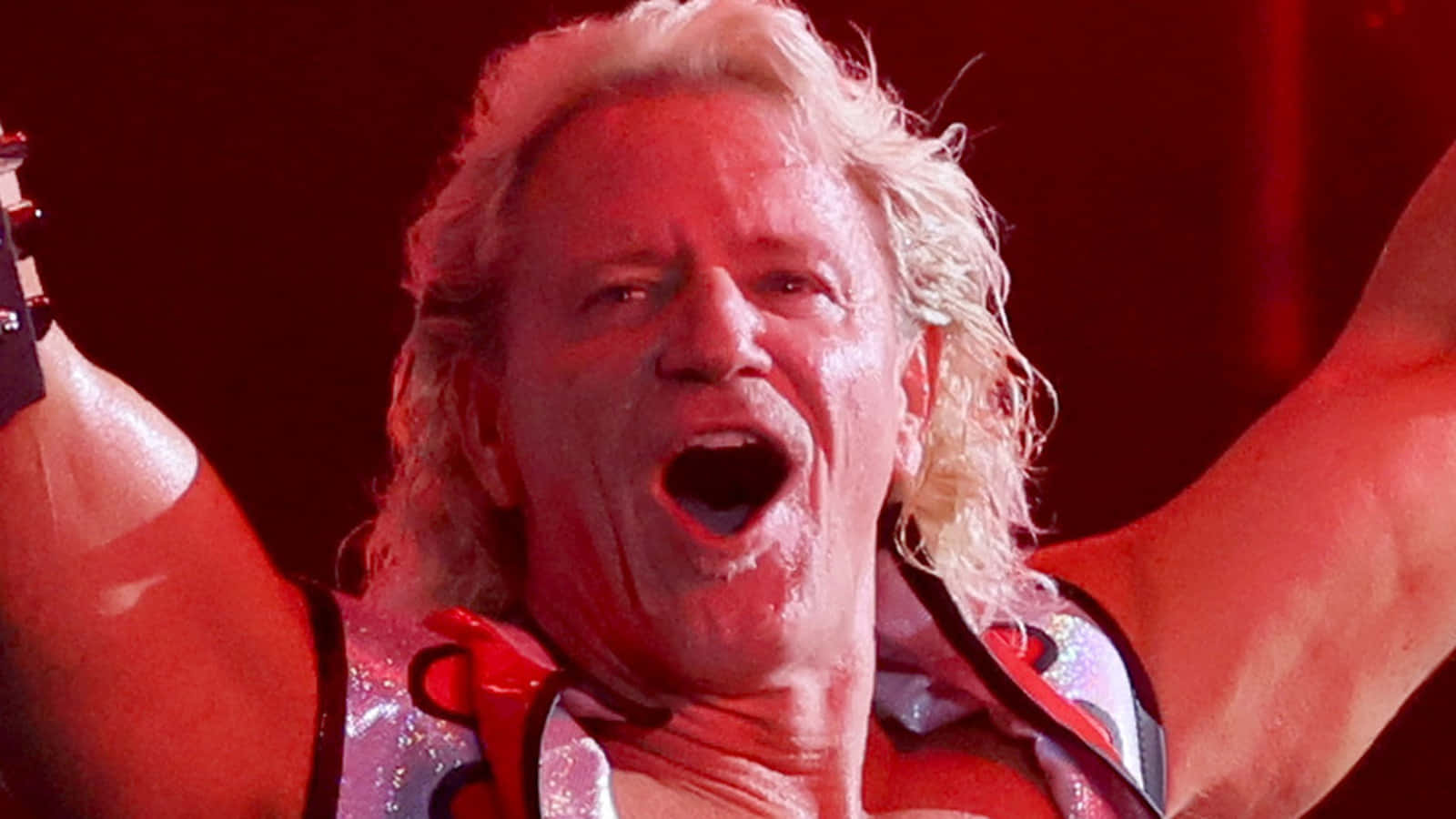 傑夫·賈雷特高興慶祝 WWE 摔角手 壁紙 — 一名擁有狂野金髮的摔角手在紅色舞台燈光下穿著閃亮服裝勝利地尖叫。