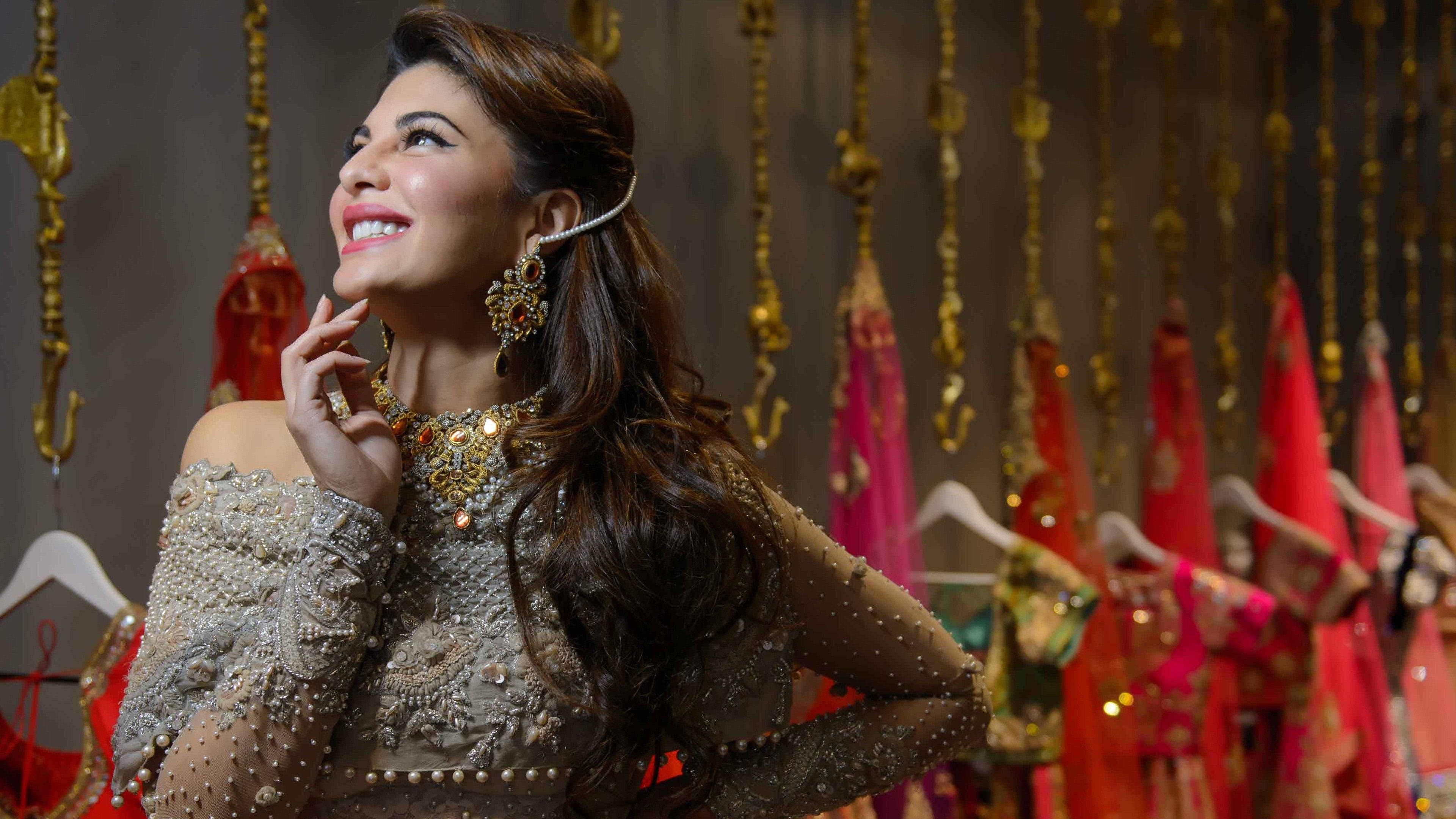 傑奎琳·費南德斯 聚光燈下 壁紙 — 一位光彩照人的女性，佩戴華麗的金飾和裝飾服飾，在懸掛的傳統服裝中向上微笑。