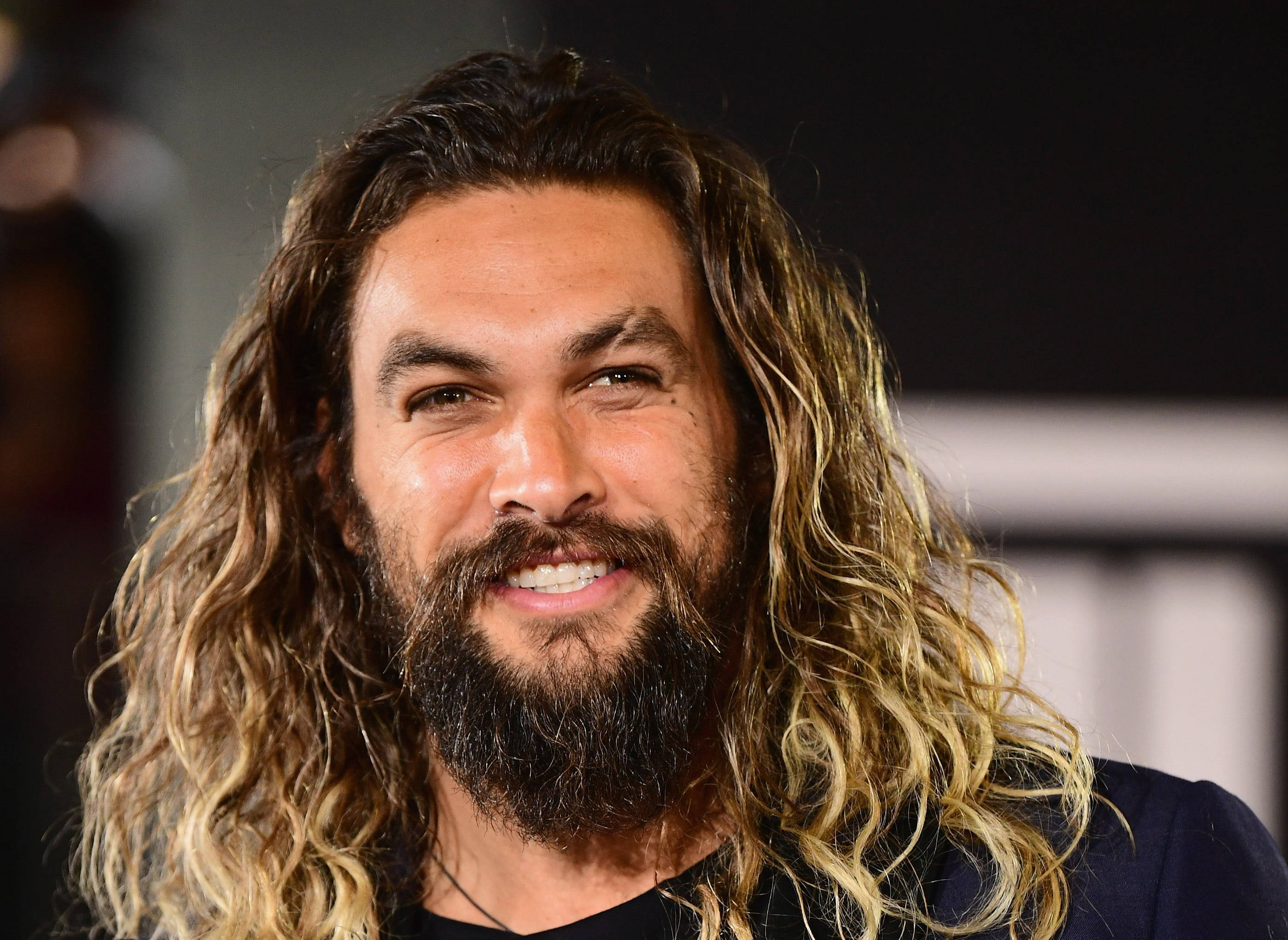 傑森·莫莫亞微笑 壁紙 — 演員積遜莫瑪（Jason Momoa）留著長長的波浪形金髮和濃密的鬍鬚，在模糊的背景前溫暖地微笑。