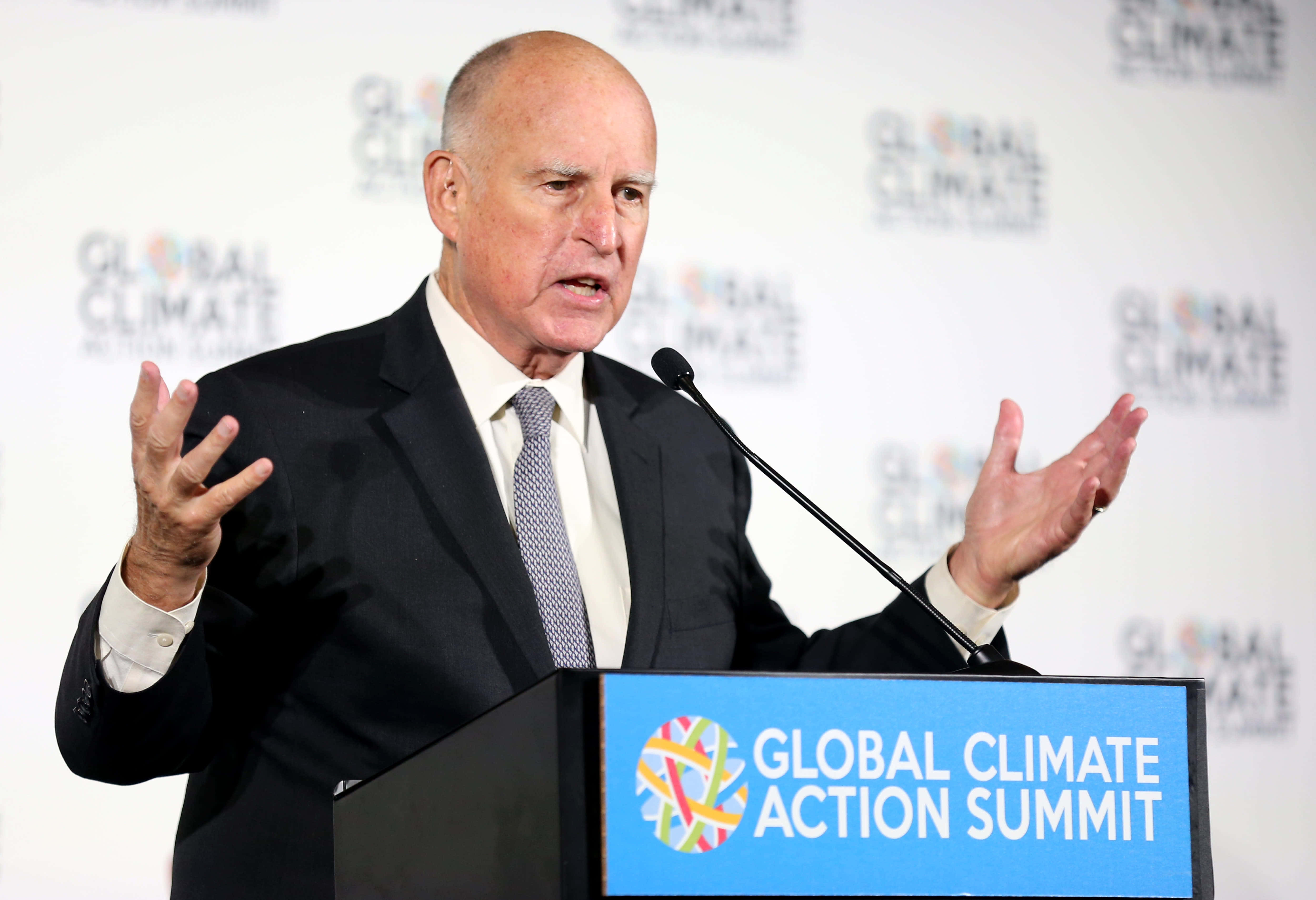 傑瑞·布朗與社區交流 壁紙 — 一名禿頭男子身穿深色西裝，在標有 Global Climate Action Summit 的講台上熱情地做手勢發言。
