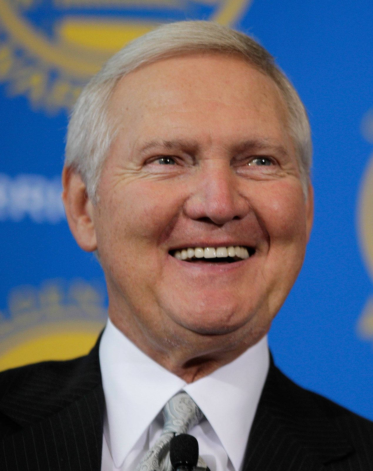 傑瑞·韋斯特 微笑 領取 NBA 經理人年度獎 壁紙 — 一位微笑的老年男士穿著西裝，背景為藍色並帶有黃色標誌的特寫。