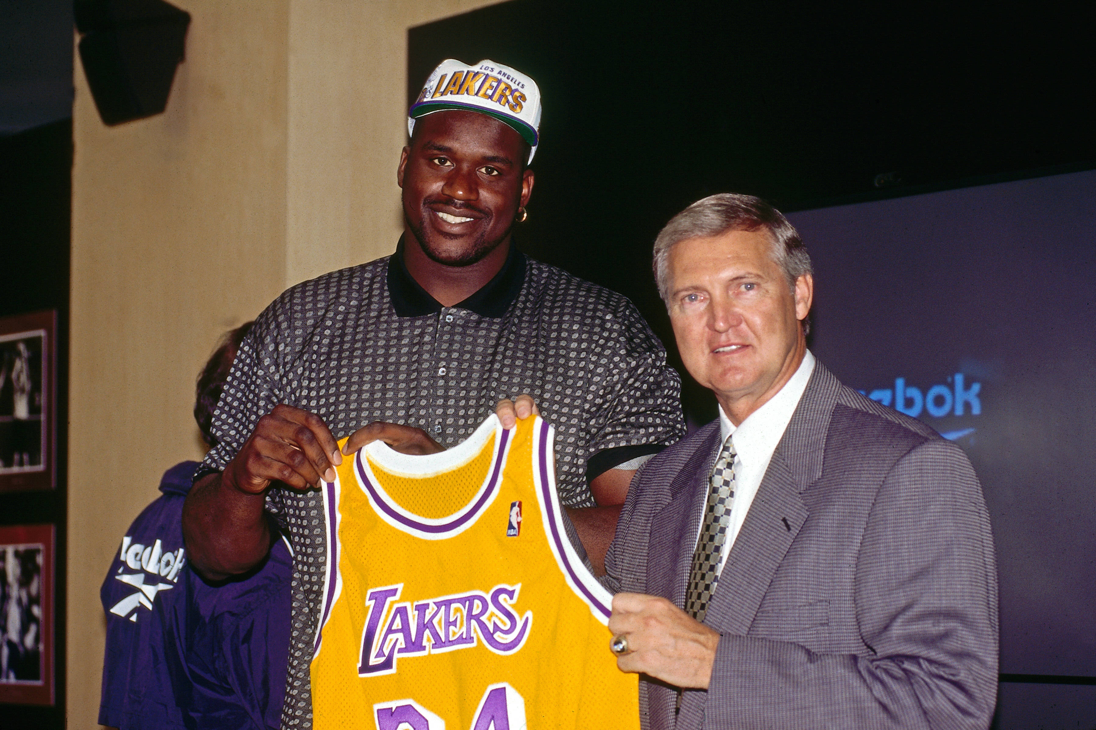 傑瑞·韋斯特歡迎沙奎爾·奧尼爾加盟湖人隊 壁紙 — Shaquille O'Neal 笑容滿面，手持一件黃色湖人隊34號球衣，一名西裝男子與他一同拿著。
