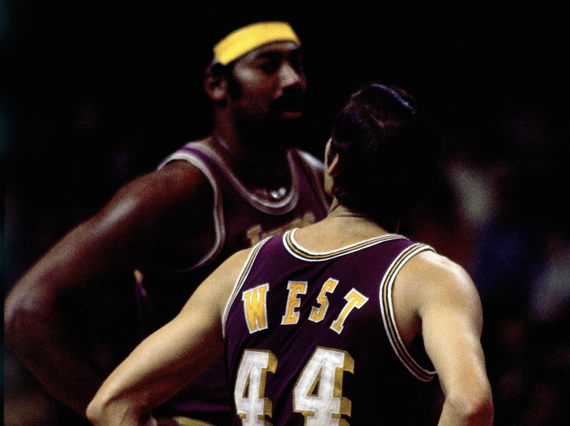 傑瑞·韋斯特與威爾特·張伯倫 NBA史上最強雙人組合 壁紙 — 兩名身穿紫色和金色球衣的籃球運動員在場館燈光下激烈對峙。