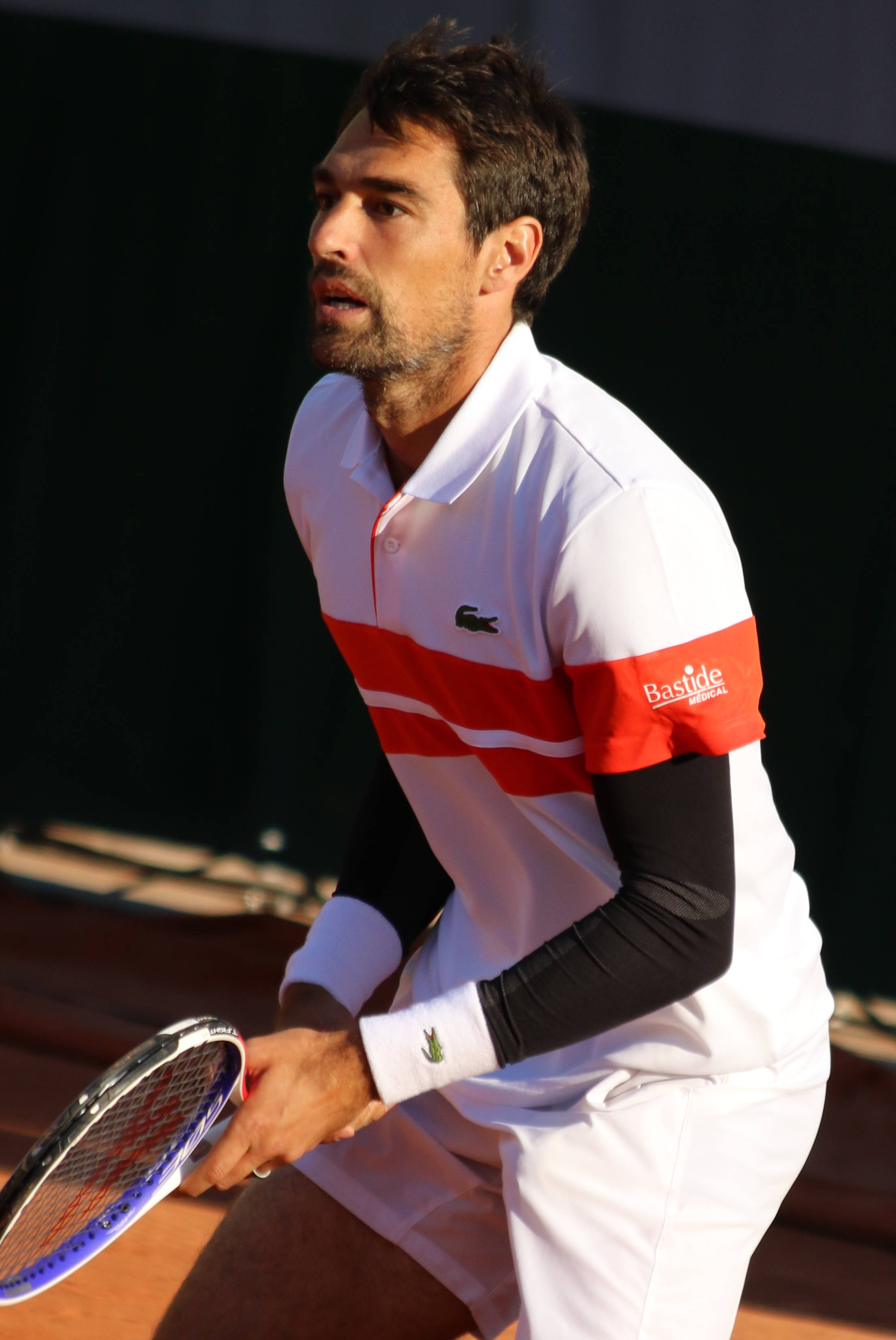 傑瑞米·夏迪，Lacoste贊助網球選手 壁紙 — 一位專注的男性網球選手，身穿白色和紅色裝備，在陽光普照的紅土球場上握緊球拍。
