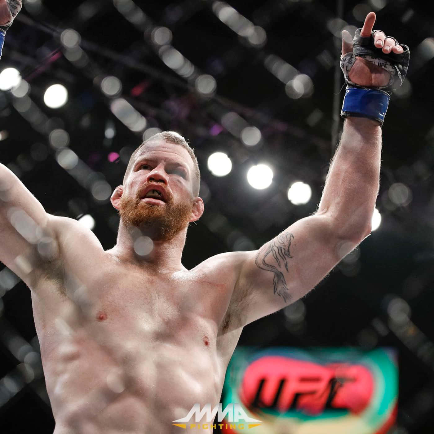 傑瑞米·霍恩UFC比賽夜結果 壁紙 — 一位留鬍鬚的勝利MMA拳手在燈光明亮的八角籠內高舉戴手套的拳頭慶祝勝利。