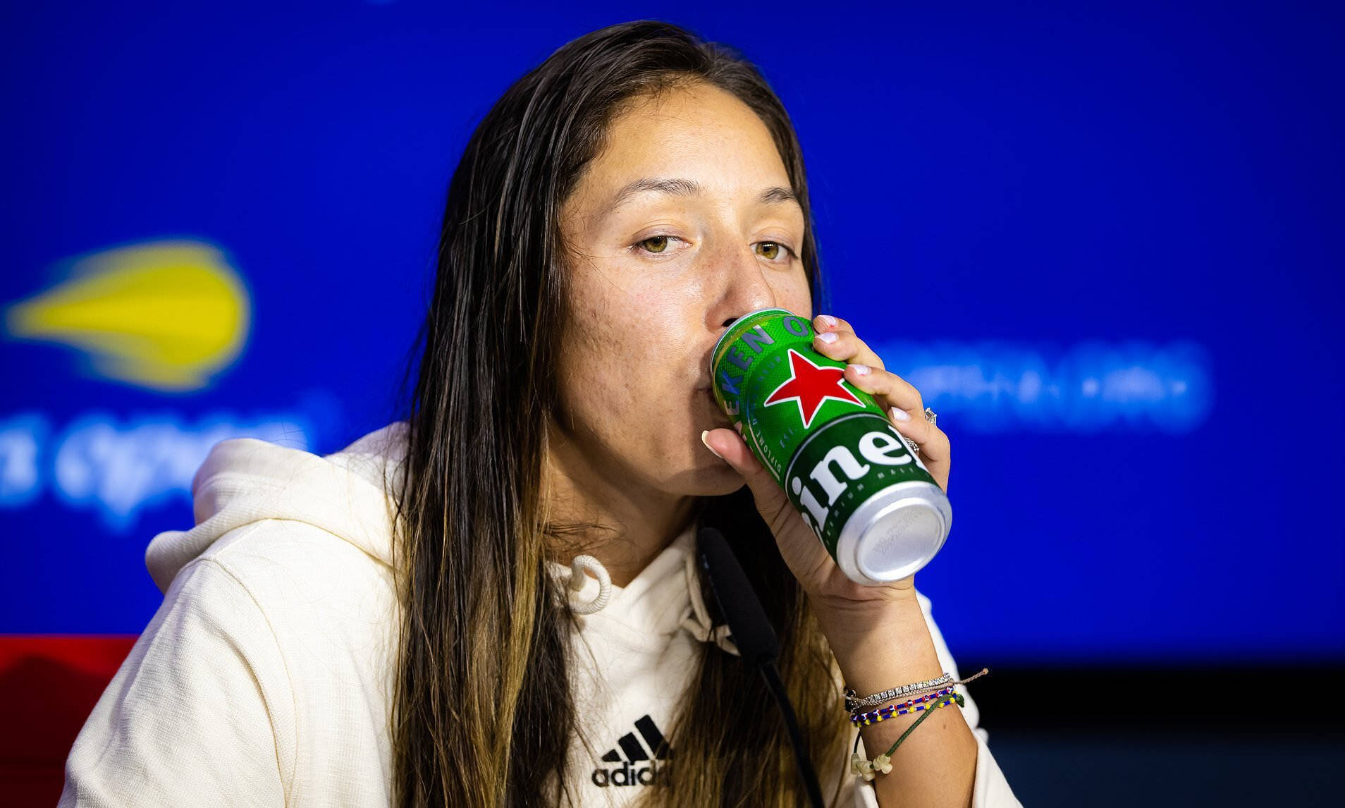 傑西卡·佩古拉飲啤酒 壁紙 — 一位身穿白色連帽衫的女性在藍色背景前喝著綠色喜力啤酒罐，背景有模糊的標誌。