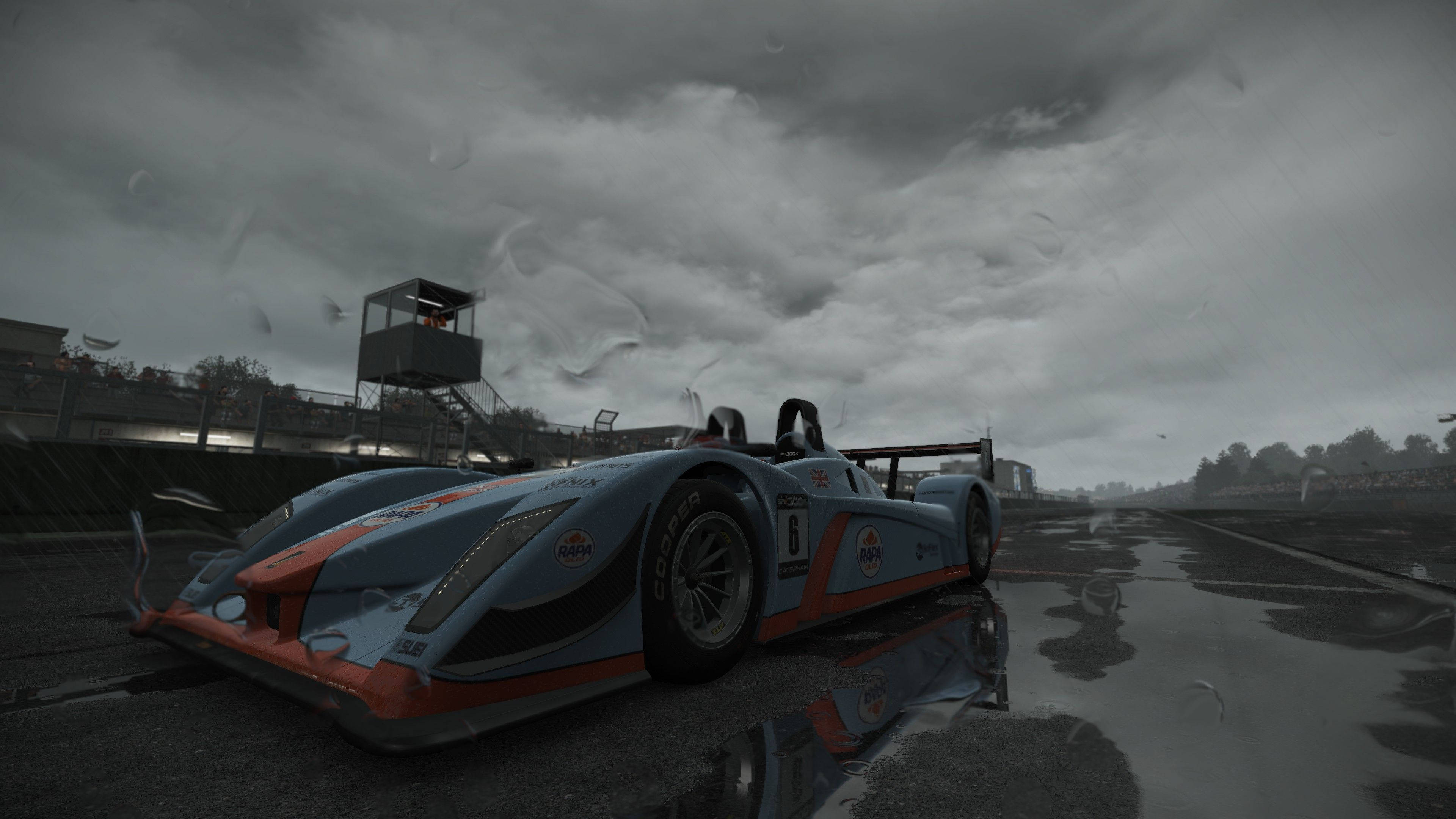 傲視群雄：Project Cars 4K 中的奧迪 R8 壁紙 — 一架流線型的藍色和橙色賽車在風暴般的灰色天空下，靜止在潮濕的賽道上，鏡頭上可見雨滴。