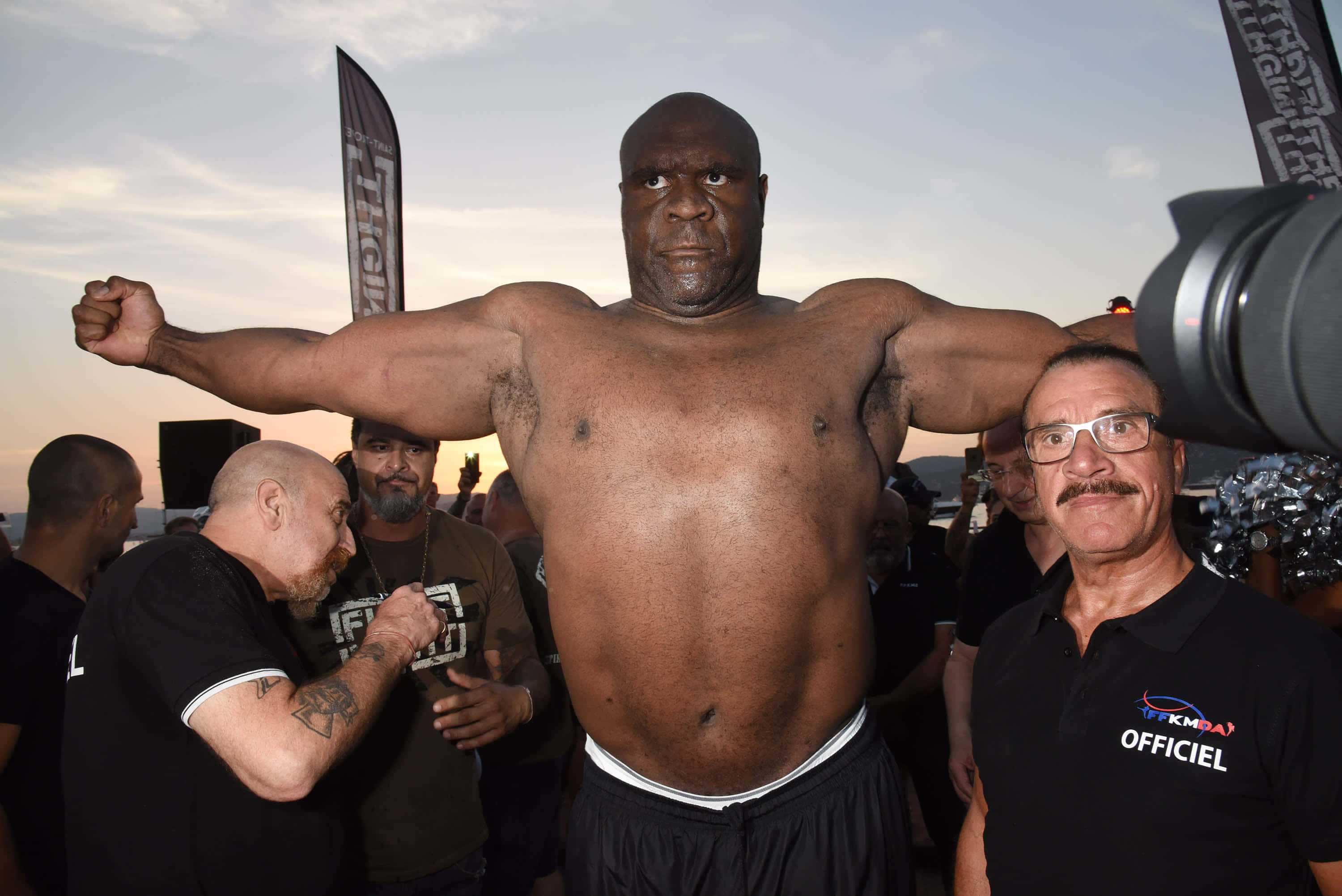 傳奇 Bob Sapp 的賽前會議 壁紙 — 一名肌肉發達的男子在戶外屈起手臂，官員在日落時分的公開活動中為他稱重。