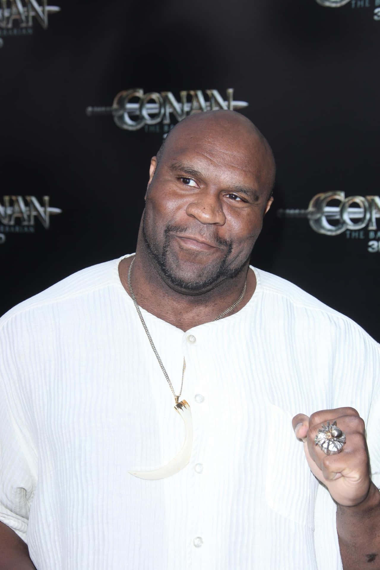 傳奇 Bob Sapp 肖像 壁紙 — 一個光頭男人穿著白色鈕扣襯衫，在「王者之劍」活動中微笑著舉起一個大型裝飾戒指。