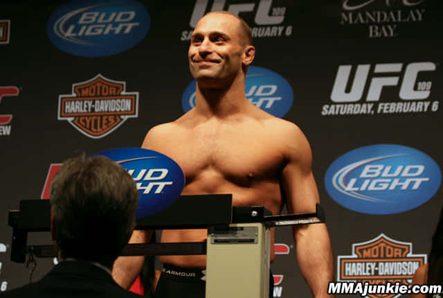 光頭MMA拳手在UFC稱重時微笑，身後有贊助商標誌。