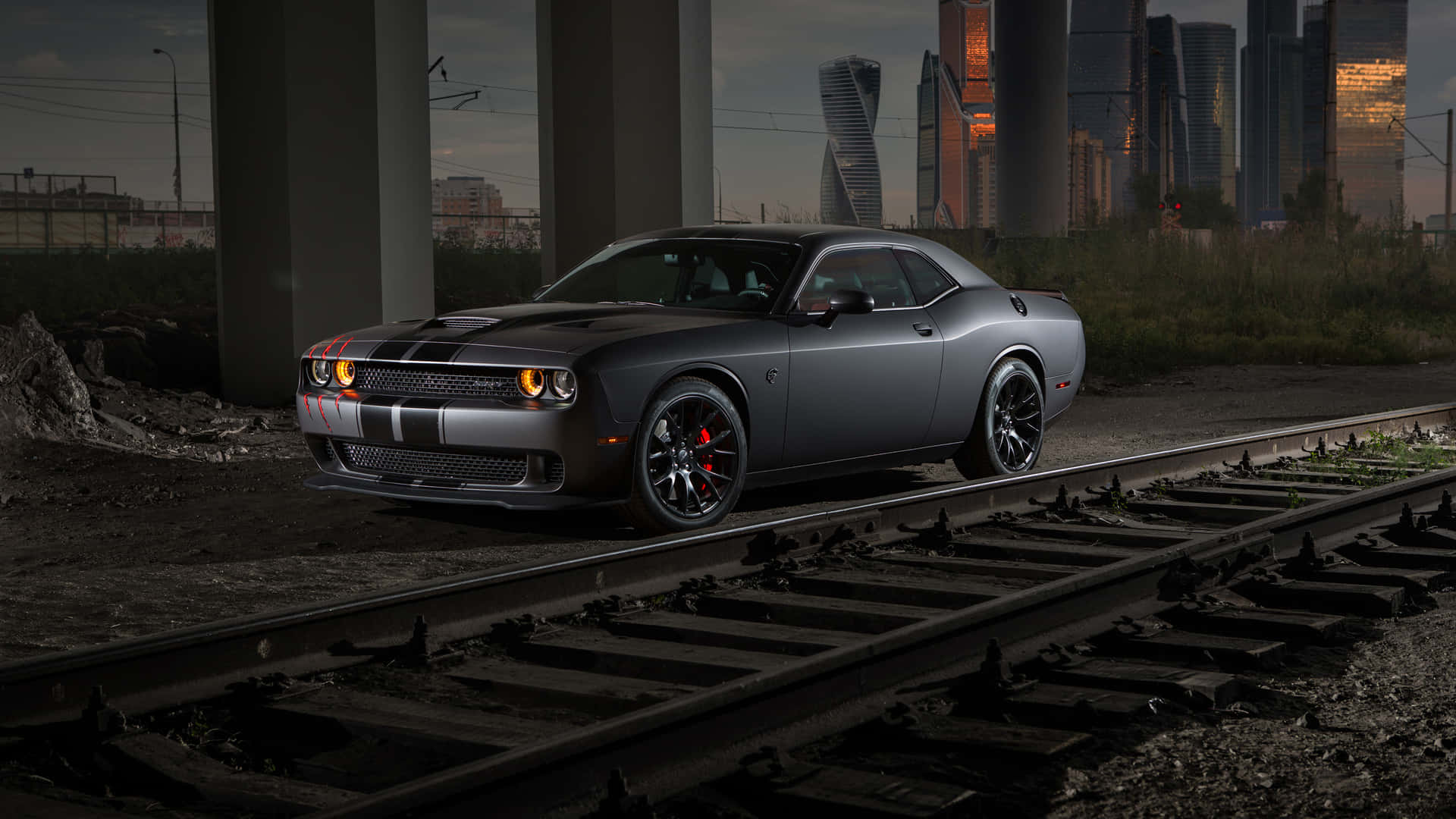 灰色Dodge Challenger停喺廢棄火車軌上，背景係黃昏城市天際線。