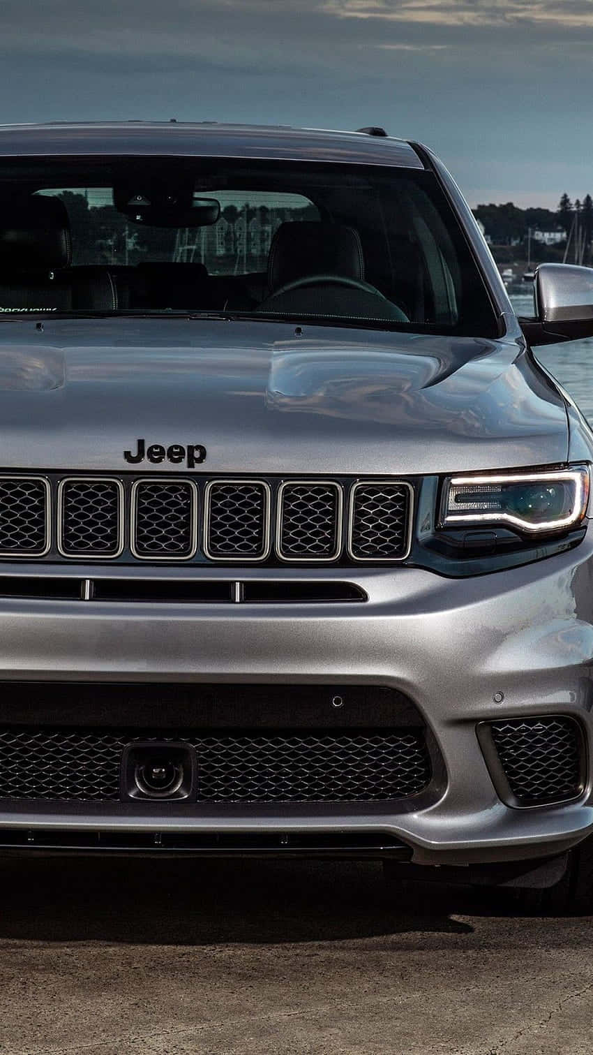 銀色Jeep SUV正面停喺水邊，背景係多雲嘅天空。