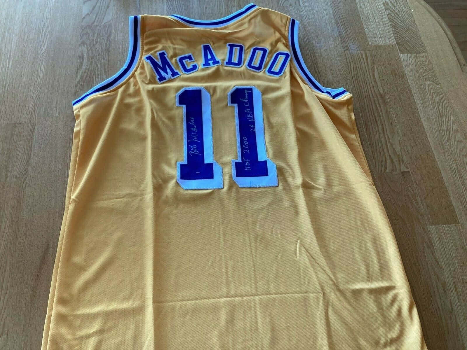 傳奇NBA球星鮑勃·麥道奧身穿11號湖人隊球衣 壁紙 — 一件金色籃球球衣，上面有藍色 'McADOO' 和號碼 '11'，平放在木地板上，簽名並刻有 'HOF 2000' 和 'NBA Champ'。