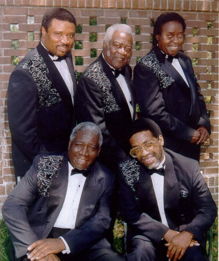 傳奇R&B樂團 The Drifters 壁紙 — 五位微笑的黑人男性穿著華麗的黑色燕尾服，自信地以帶有綠色瓷磚的磚牆為背景擺姿勢。